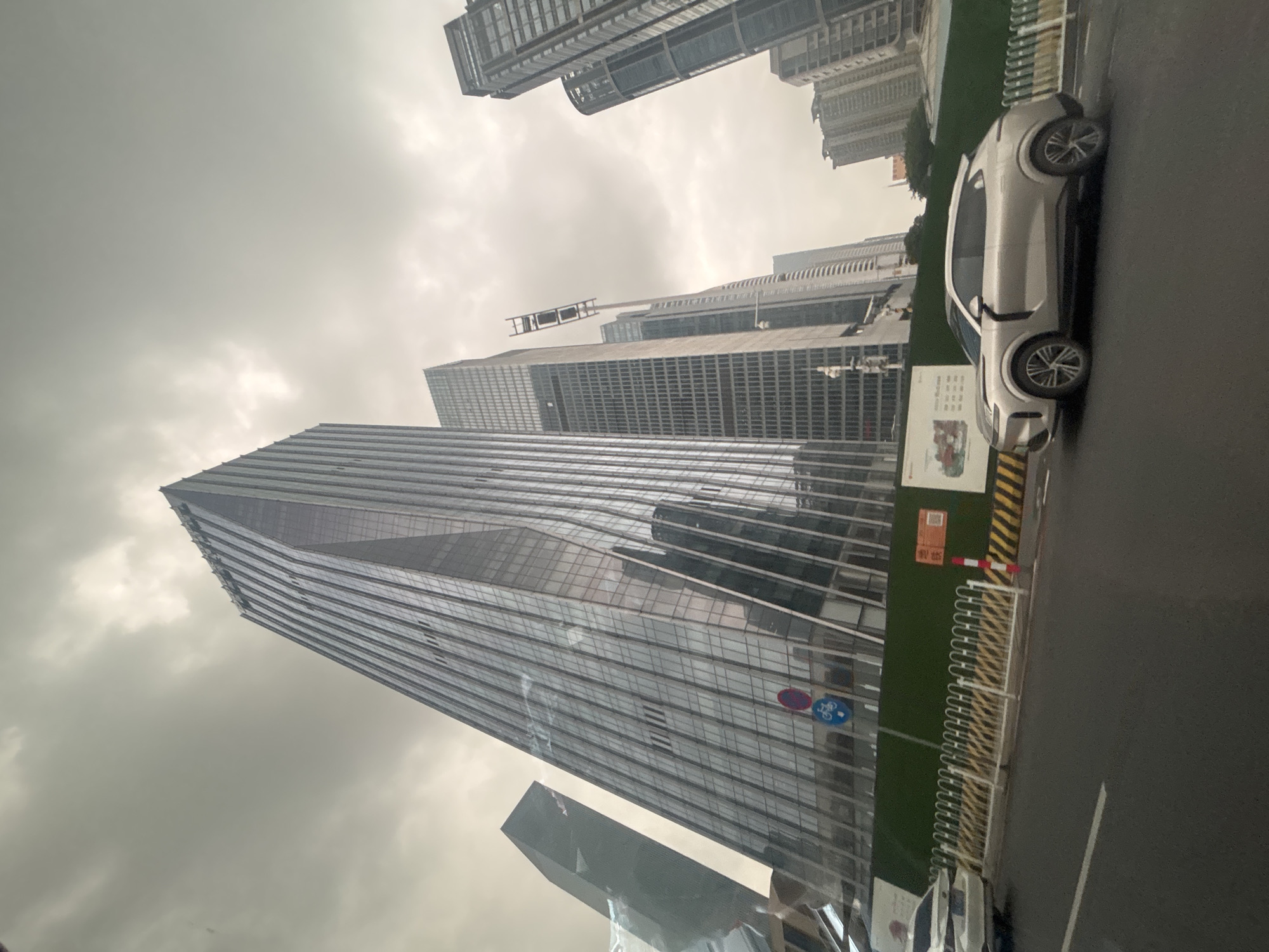 The image captures a street-level view of modern skyscrapers in Tianhe District, China, under a cloudy sky. In the foreground, a silver SUV is parked on the right side of the road, facing left. To the left of the SUV, a white sedan is partially visible. The road is asphalt with white lane markings. A green construction fence runs along the sidewalk, behind which several signs are posted, including one with Chinese characters and a QR code. A row of yellow and white striped barriers is visible along the fence. The dominant features are the tall, sleek glass-fronted buildings, reflecting the overcast sky. A traffic sign with a red circle and a blue bicycle symbol is affixed to one of the buildings. The overall impression is one of urban development and a slightly muted, perhaps damp, atmosphere.