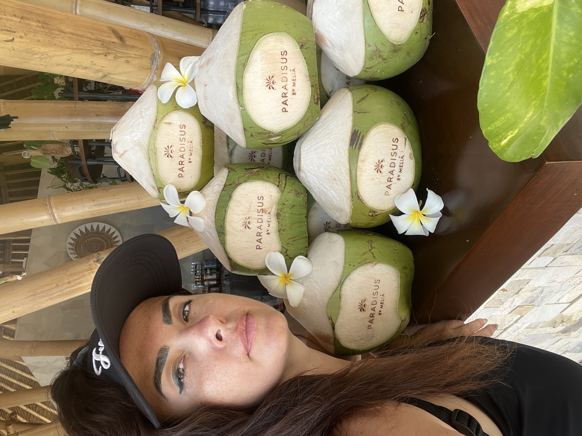 A woman wearing a black visor and black tank top is posing for a selfie in front of a display of green coconuts with white labels. The coconuts are stacked in a pyramid formation, and each label features the text "PARADISUS BY MELIA" along with a palm tree logo. White flowers are placed on top of some of the coconuts. The background consists of large bamboo poles, thatched roofing, and a glimpse of an indoor setting with tables and chairs. The scene appears to be in Kuta Selatan, Indonesia, likely at a resort or restaurant.