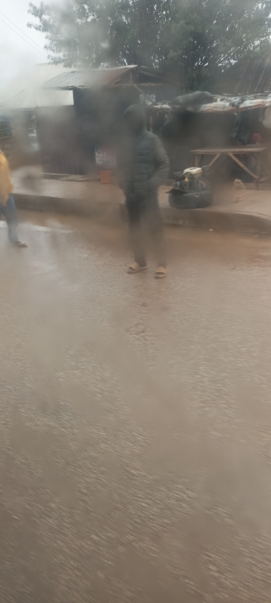 The image displays a blurry, rain-affected street scene, likely captured through a wet or fogged lens. The foreground features a wet road surface, indicating recent or ongoing rain.
Two individuals are discernible. One person stands centrally in the frame, wearing a dark, possibly puffy, jacket and light-colored sandals. To the left, another person is partially visible, dressed in a yellow top and blue trousers.
In the background, several informal structures, likely small shops or dwellings, line the street. These buildings appear to have corrugated iron roofs. Trees are visible further behind the structures. On the right side of the image, near the centrally-placed individual, a small engine or generator is resting on a tire, next to a wooden table or bench. An orange bucket is also visible in front of one of the structures.
The overall atmosphere suggests overcast and rainy weather conditions. Faint text "PCG" can be seen on one of the structures. Another partially obscured sign appears to have the word "SERVICE". The scene depicts a common street setting in what appears to be a semi-urban or rural area, consistent with Bukuru, Nigeria, during inclement weather. Due to the significant blur, specific facial expressions or detailed activities beyond people standing are not discernible.
