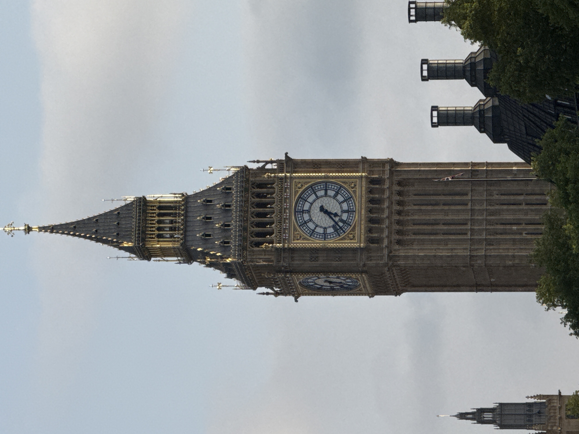 The image features the Elizabeth Tower, commonly known as Big Ben, in London, United Kingdom. The tower is prominently displayed against a backdrop of a grey sky. A portion of the Houses of Parliament is visible to the right, partially obscured by trees. Another tower appears distantly in the bottom right corner. The clock face of Big Ben is visible.