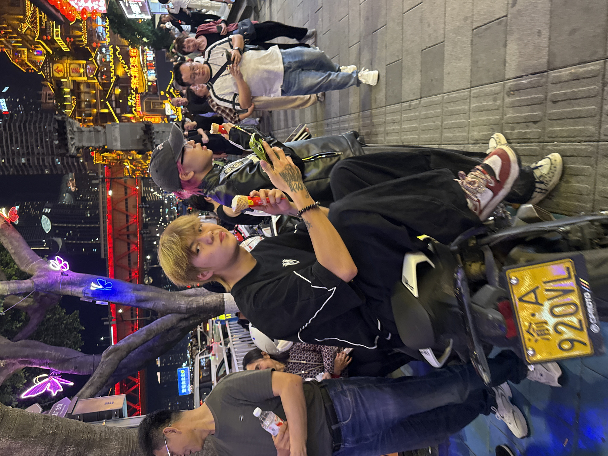 The image depicts a crowded street scene in Yuzhong District, Chongqing, China, at night. Artificial lights illuminate the area with multiple buildings and pedestrian traffic.
People:
The picture includes many people. The main subject is a blonde male seated on a vehicle. Another individual next to him is holding a phone. An older man is drinking from a bottle.
Subjects:
The primary subjects are individuals in the foreground.
Objects:
Objects include clothing (jackets, jeans, sneakers), personal accessories, phones, ice cream cones, a motorcycle or scooter, and a license plate. A bottle of water is also visible. The license plate displays "渝 A 920VL", corresponding to Chongqing.
Actions:
People are observed eating, using cell phones, standing, and sitting.
Interactions:
Individuals appear to be interacting with each other.
Environment:
The setting is an urban street at night. Decorated buildings and trees with artificial lighting suggest a tourist area or commercial district. There is a wet sheen to the walking path. The overall atmosphere is festive with lights.