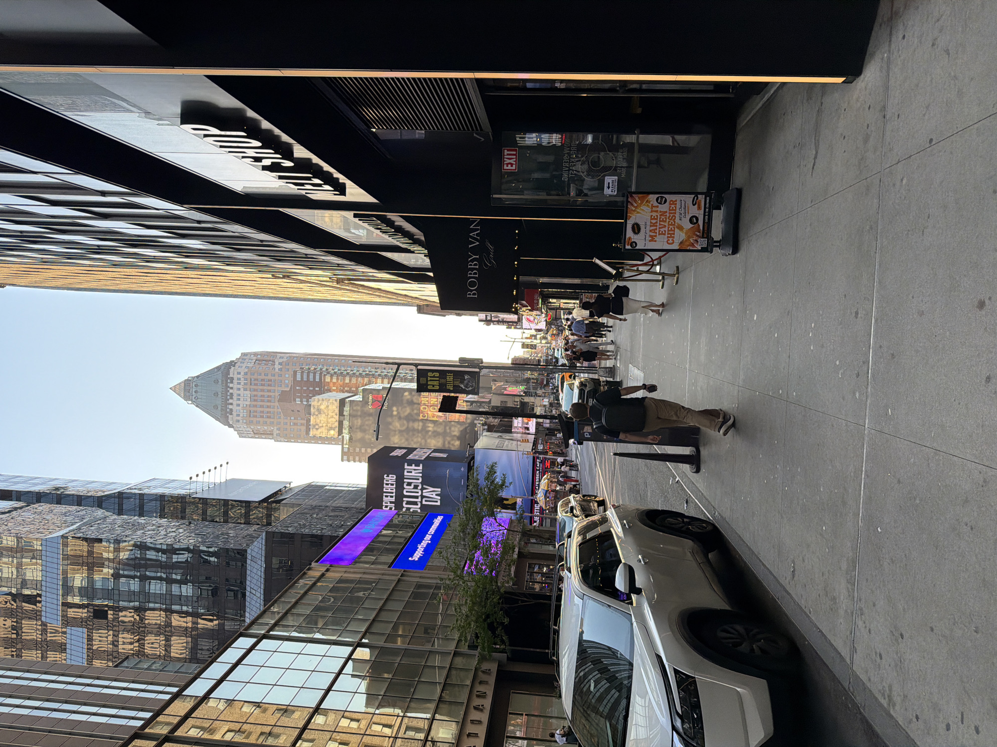 The image depicts a street scene in New York City, United States of America. The perspective is from street level, looking down a sidewalk. The street is lined with buildings, and there are several people walking. The sidewalk on the right of the image is where the Bobby Van Grill restaurant is situated, with an exit sign visible. The city's recognizable buildings and landmarks appear in the background. The electronic billboards display a Spielberg production and a message that reads, "SPIELBERG...SCLOSURE DAY...Supporting our communities." A silver car is parked along the curb on the right side of the image.