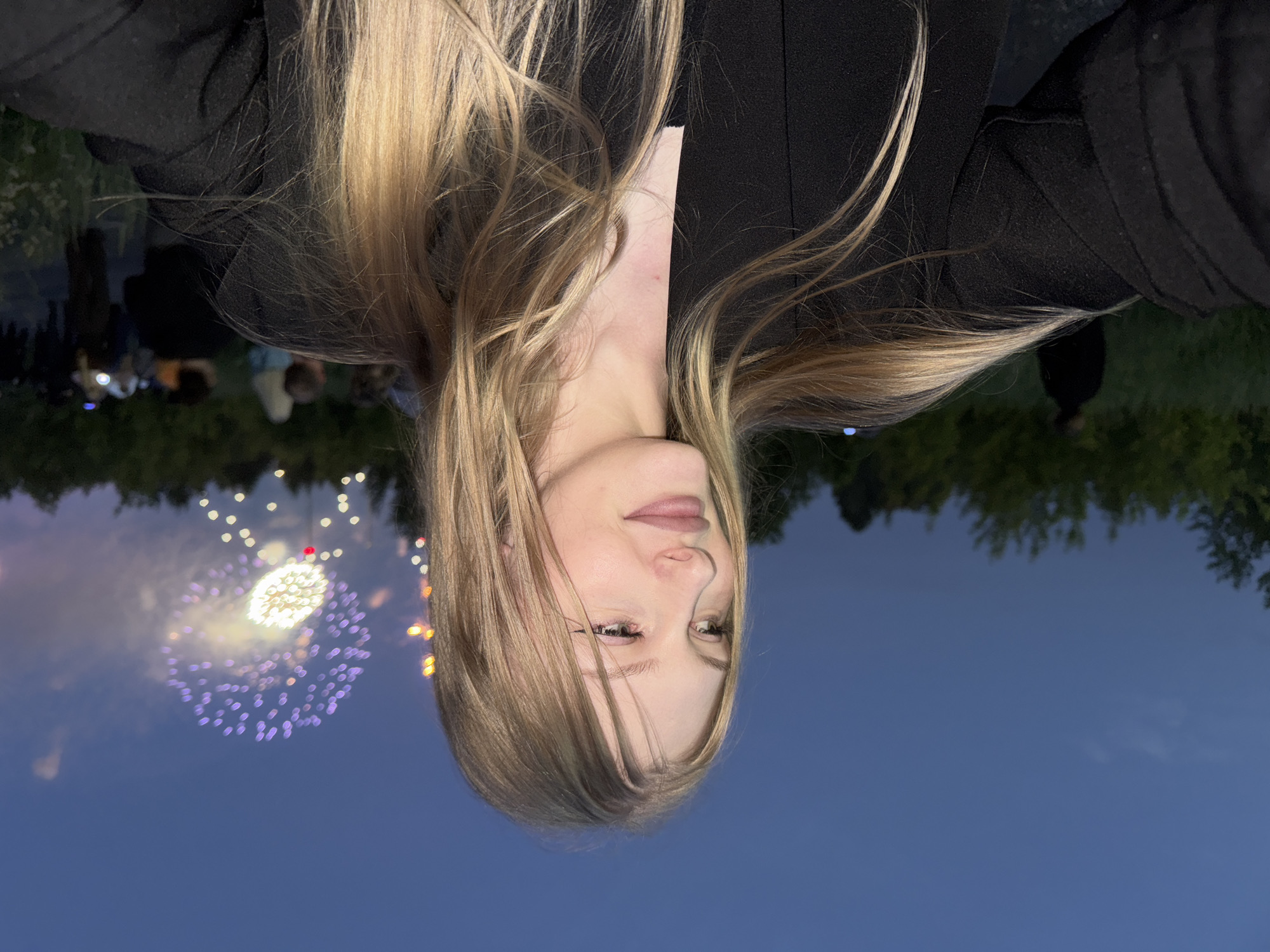 A young woman with long blonde hair is the main subject of this photograph, taken outdoors at dusk. She is wearing a black jacket and a black top. Her face is turned to her left, looking off into the distance with a slight smile. In the background, fireworks are visible in the dark blue sky. A line of trees is silhouetted against the sky on the horizon. Several other people are visible in the background, some appearing to be watching the fireworks. The ground appears to be grassy. The exact location in Perm, Russia cannot be determined from the image.