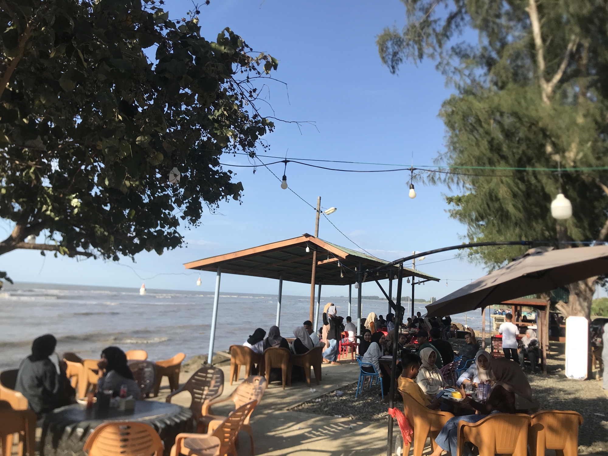 The image shows an outdoor seating area near a body of water, likely a beach, in Lancok, Indonesia. Several people are seated at tables, some under a covered structure with hanging light bulbs, and others under umbrellas. The scene appears relaxed, with people eating and socializing. The environment includes trees, the sea, and a clear sky. The chairs and tables are made of plastic and are arranged in a casual manner. There is a concrete area under the seating arrangement with some debris. The sea is visible in the background with light waves.