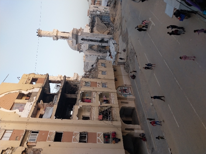 The image depicts an urban street scene showing the aftermath of significant structural damage to buildings, with people on the street and a mosque in the background. The photo is taken during daytime under a clear, light blue sky.
On the left side of the frame, several multi-story residential buildings are severely damaged. The facades are largely a light beige or yellowish color, with some patterned brown and white brickwork. Many sections of these buildings are blown out or collapsed, exposing their interior rooms, concrete slabs, and reinforcing rebar. Some areas appear charred and blackened, indicating fire damage. Broken windows and gaping holes are common. In some exposed rooms or balconies, items resembling hanging laundry (red and white fabrics) are visible. Further back and to the left, another partially destroyed building stands.
In the mid-ground, slightly to the right of the center, a mosque with a tall, white minaret is visible. The minaret is cylindrical, featuring decorative elements and a balcony near its top, culminating in a pointed finial. The mosque's dome is also visible next to the minaret. Both the minaret and dome appear to be in relatively good condition compared to the surrounding residential structures, though parts of buildings immediately around the mosque also show signs of damage.
The right side of the image features a wide, paved street that runs diagonally from the lower left to the upper right. The street has distinct white markings, possibly lane dividers or parking lines. A dirt or unpaved shoulder borders the street on its left side.
Various people, a mix of adults and children, are scattered across the street and its vicinity. They appear to be walking or standing in small groups. One individual near the center-right is wearing a red shirt and blue pants. Another child in a pink outfit is visible towards the far right. Most other figures are dressed in darker clothing. Their postures appear casual, suggesting everyday activities despite the damaged surroundings. In the far upper right, near what looks like a makeshift tent or shelter, more figures are gathered.
The overall scene conveys a stark contrast between daily life continuing on the street and the widespread destruction of residential buildings, with a prominent religious structure standing largely intact amidst the devastation. There is no visible text in the image.