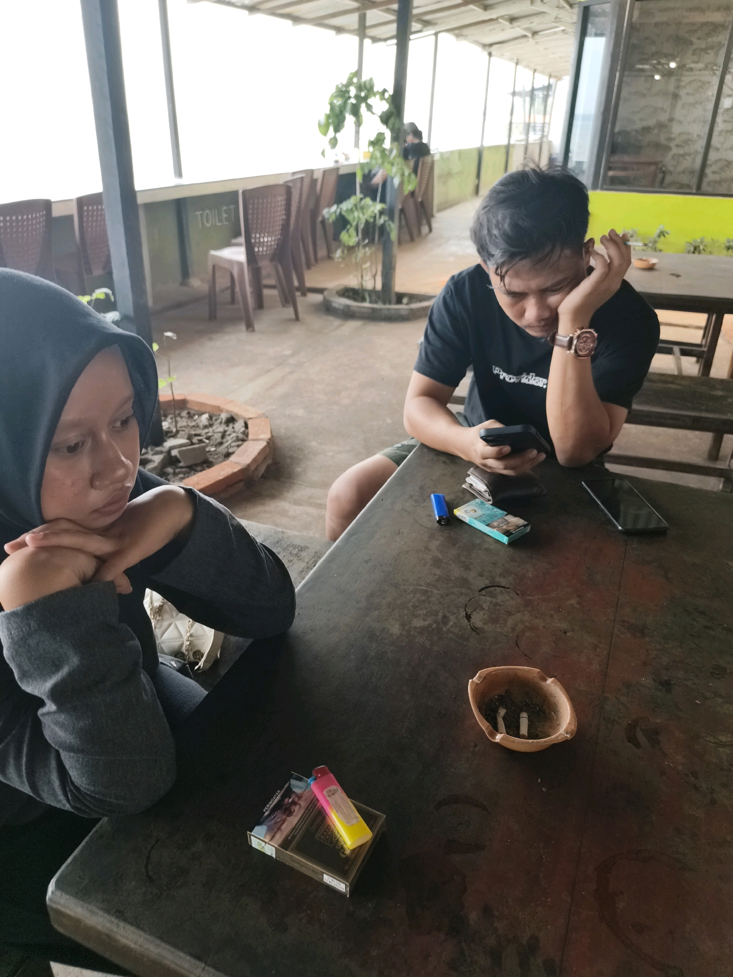The image depicts two individuals seated at a rough wooden table, likely in an outdoor dining or seating area. The person on the left, wearing a black hijab and grey hoodie, rests their chin on their folded arms, looking downwards with a contemplative expression. To the right, a man in a black t-shirt leans forward, one hand supporting his head while he looks at his mobile phone. He wears a wristwatch on his left wrist.
On the table, various objects are visible: a pack of cigarettes, a lighter, a smartphone, a wallet, and an ashtray containing cigarette butts. A second pack of cigarettes with a pink and yellow lighter rests closer to the foreground. The environment appears to be casual, possibly a roadside cafe or a similar establishment. In the background, there are rows of brown plastic chairs and a sign that reads "TOILET". The overall lighting suggests it is daytime. The mention of Ketapang, Indonesia, suggests a regional context for this scene.