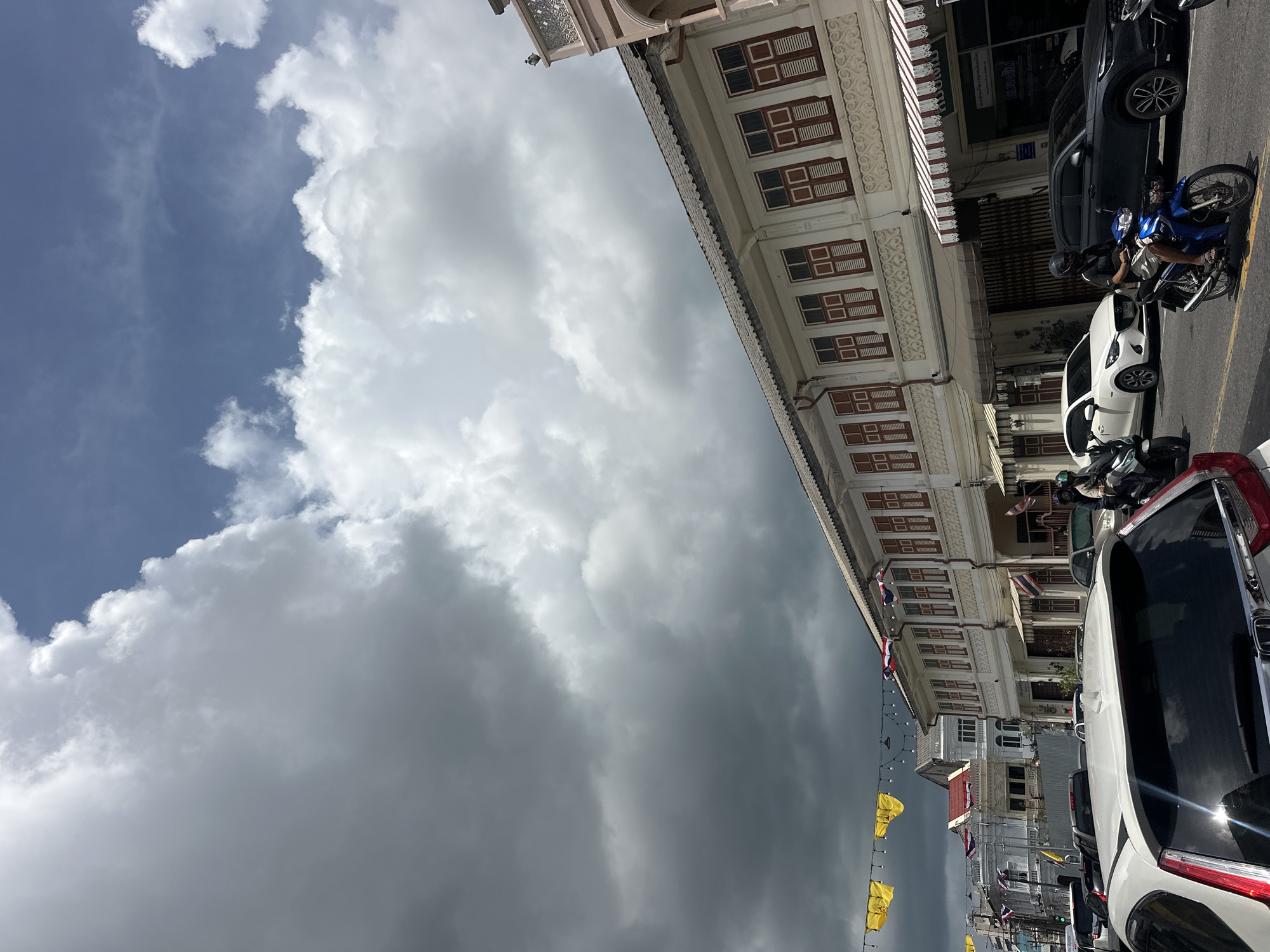 The image depicts a street scene in Phuket City Municipality, Thailand, under a dramatic sky with heavy, dark clouds contrasting with patches of blue sky and bright white cumulus clouds. The architecture suggests a historic district, with buildings featuring white facades, numerous shuttered windows with red frames, and decorative architectural details. Yellow and triangular flags are strung across the street, along with smaller Thai flags on some buildings. Traffic is present, including several cars and motorcycles. One motorcycle rider, wearing a helmet and dark clothing, is visible in the foreground. The lighting indicates it is daytime, with the sun partially obscured by clouds.