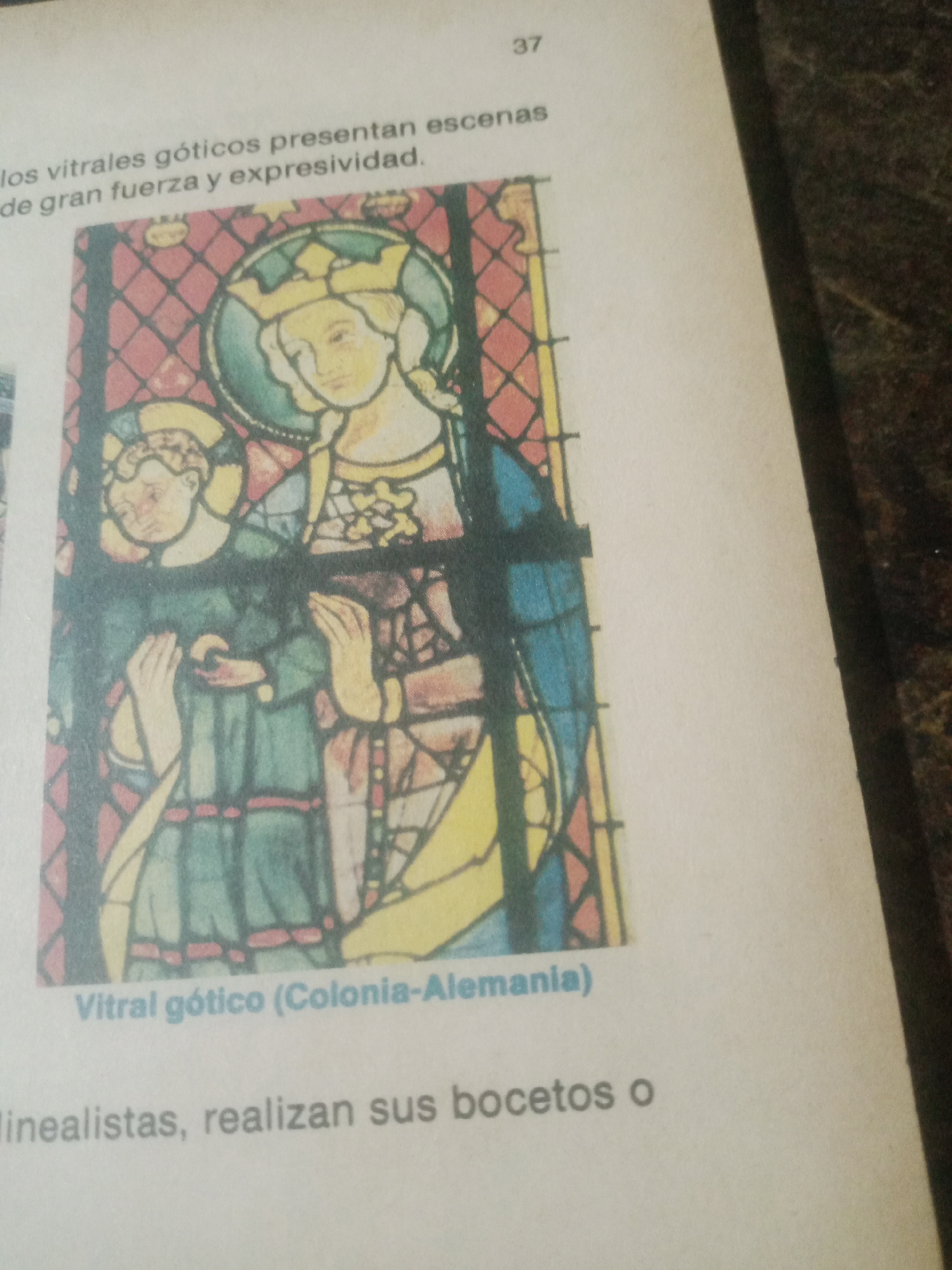 The image displays a page from a book or document featuring a stained-glass window illustration and accompanying text. The stained-glass image depicts a religious scene, likely of a Madonna and child, with figures rendered in a stylized, medieval fashion. The dominant colors are blue, green, yellow, and red, set against a background of diamond-shaped patterns. Below the illustration, text in Spanish identifies the artwork as "Vitral gótico (Colonia-Alemania)," which translates to "Gothic stained glass (Cologne-Germany)." Further text below this reads, "linealistas, realizan sus bocetos o," implying a discussion about stylistic approaches and the creation of sketches. The page also has the number "37" in the upper right corner. The image quality is slightly blurry, suggesting it was captured with a handheld device. The context of Santa Ana, Venezuela, is not present in the visual information provided and cannot be inferred from the image or its text.