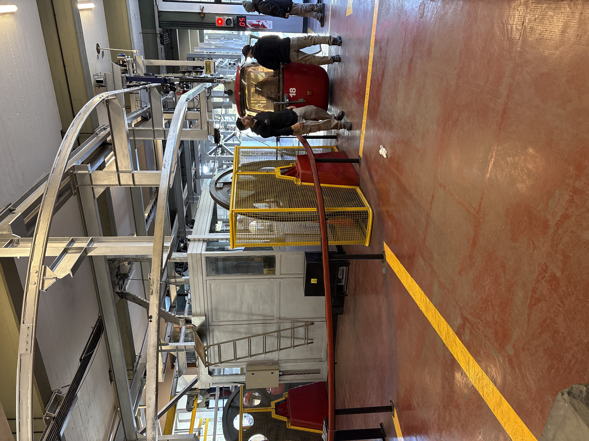 The image displays the interior of a cable car station, identified as being in San Carlos de Bariloche, Argentina. The floor is reddish-brown, marked by yellow linear demarcation strips and scuff marks. A small piece of white debris rests on the floor in the mid-ground. Overhead, a robust industrial structure of interconnected grey metal beams, tracks, and mechanical components supports the cable car system.
A red gondola, numbered "18," is positioned at a boarding point in the mid-ground. Its windows are tinted, revealing indistinct reflections of occupants or interior structure. Three adult males, consistent in appearance with station operators, are present. The male closest to the left is facing right, standing near a yellow metal mesh safety barrier protecting a large industrial wheel. Another male stands with his back partially to the camera, positioned at the open entrance of the gondola. A third male stands further right, facing left towards the gondola, next to a wall-mounted fire extinguisher and a red digital display indicating "05."
To the left and rear of the gondola, a light grey, rectangular control or utility structure stands, against which a metal ladder is leaned. An electrical box is mounted on its exterior. A black box with a yellow warning sticker is on the floor in front of this structure. An orange safety rope barrier, supported by black stanchions, spans the foreground. Natural light enters from a large opening or window on the far right side of the scene.