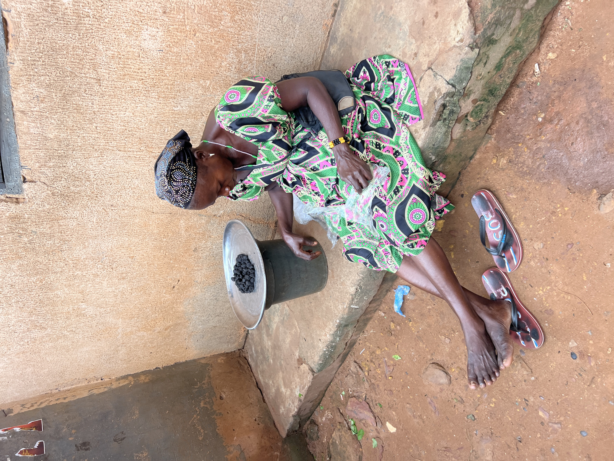 A dark-skinned woman, wearing a vibrantly patterned green, pink, and black dress and a black headscarf adorned with rhinestones, sits on a concrete ledge outside a building. Her legs are crossed, and her bare feet rest on a pair of red and white flip-flops. In front of her, to her left, is a metal basin filled with dark, granular material, possibly charcoal. She is holding a clear plastic bag in her hands, seemingly preparing to package the material. A black bag is slung across her shoulder. The environment appears to be an outdoor marketplace or street in Nkpor, Nigeria, with a rough, textured wall behind her and a reddish-brown dirt ground covered with small stones and debris. The scene is captured in daylight.