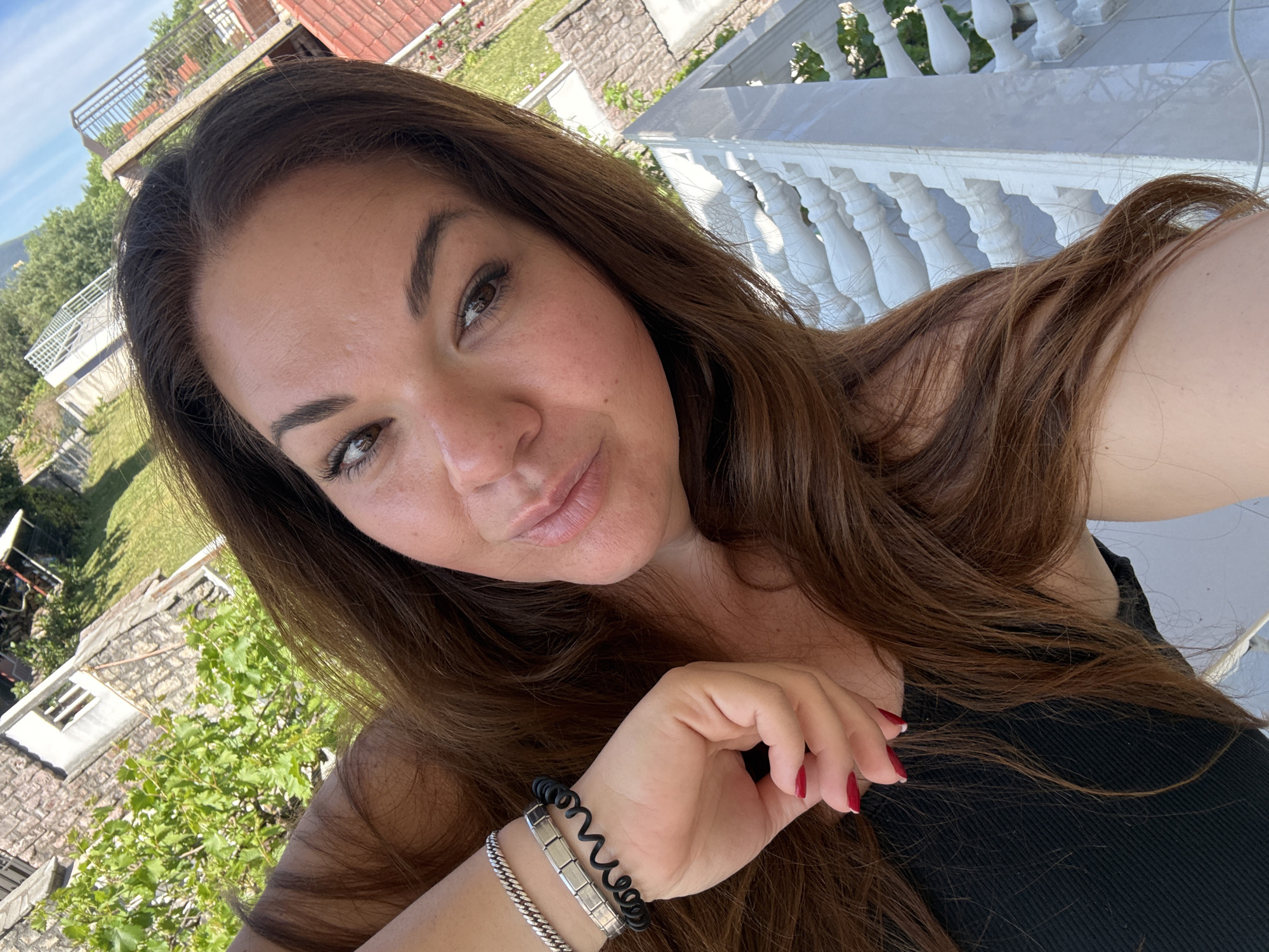 A woman with long brown hair and red nail polish is taking a selfie outdoors. She is wearing two bracelets, one silver and one black. In the background, there is a white balustrade, trees, buildings, and a partial view of the sky. Based on the architecture and landscape, the location appears to be Bijela, Montenegro.