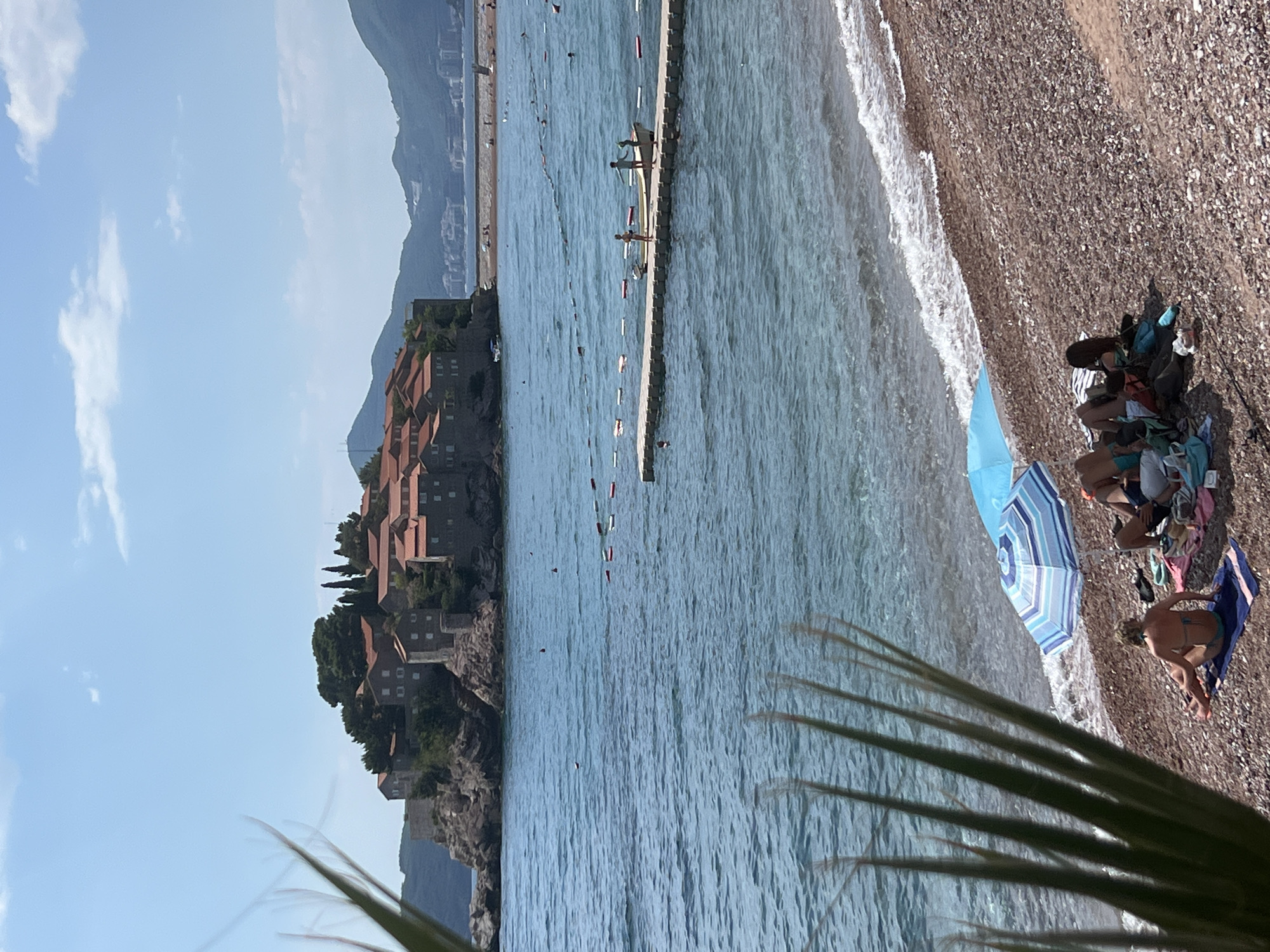 The image depicts a coastal scene in Sveti Stefan, Montenegro, showcasing a picturesque island fortified with ancient stone buildings and red-tiled roofs, situated off the mainland. The foreground features a pebbled beach with several people relaxing under beach umbrellas. One individual is seated on a towel, facing away from the camera, while others are reclined or sitting, partially obscured by umbrellas and beach accessories. The sea, a vibrant blue, stretches towards the horizon, with a floating pier extending into the water, occupied by two individuals. Red and white buoys mark a swimming area in the sea, separating it from boats. In the background, a hazy coastline with green mountains and buildings suggests a developed area. The sky is clear with a few scattered clouds, and palm fronds are visible in the left foreground, framing the scene.