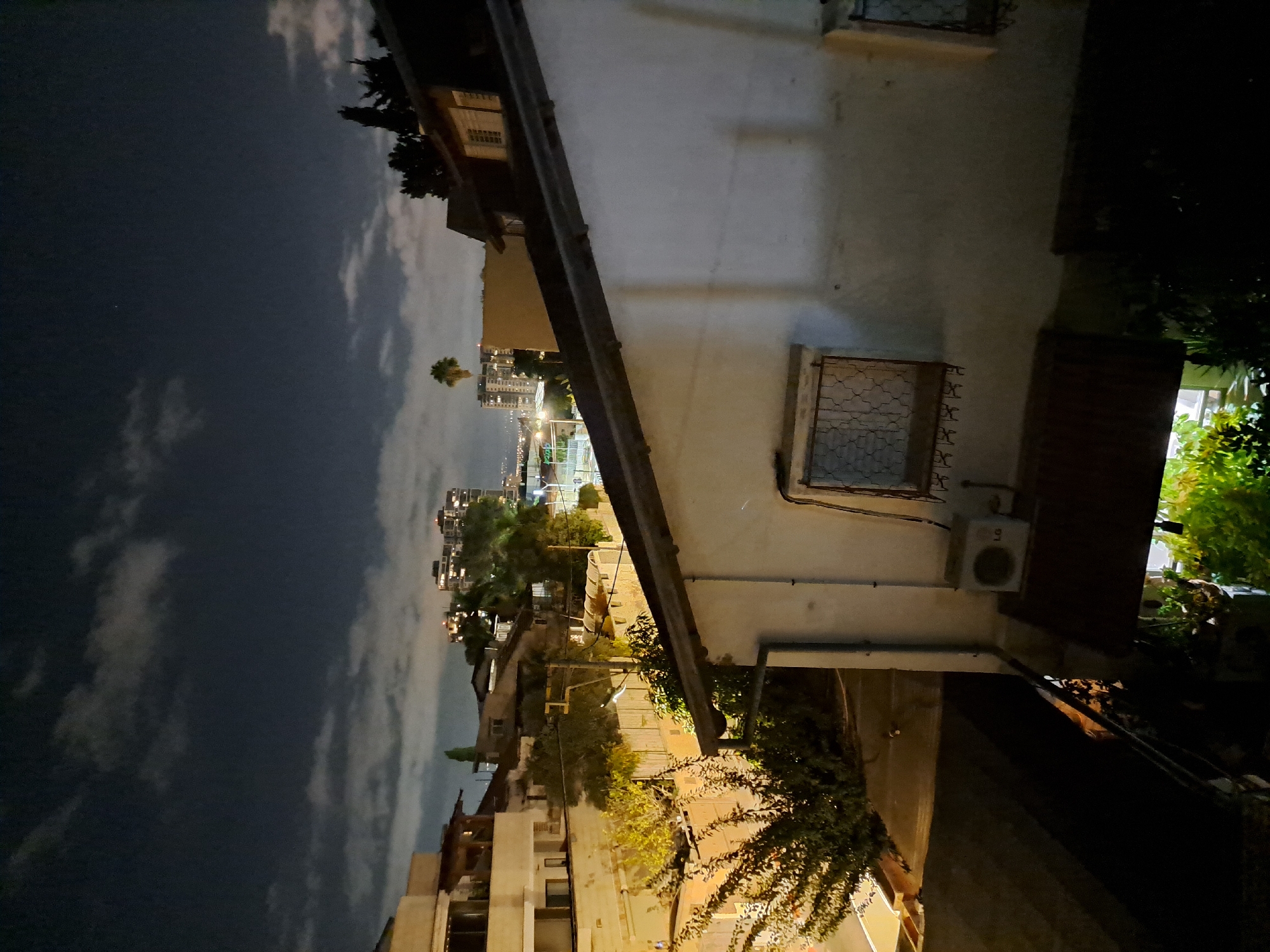 The image depicts a night view of an urban residential area in Ramat HaSharon, Israel, characterized by a mix of low-rise and mid-rise buildings under a cloudy night sky. In the foreground, a white-painted building with a sloped dark roof is visible, featuring a window with a decorative metal grate and an LG air conditioning unit mounted on its exterior wall. To the left, a dense cluster of trees with illuminated foliage suggests artificial lighting.
Further back, a complex of residential buildings with balconies and windows can be seen. Among these, a palm tree stands out against the sky. In the distance, a cluster of taller buildings with illuminated windows indicates a more densely populated urban center, possibly a commercial or business district, with streetlights visible. The overall atmosphere is one of a quiet residential neighborhood at night, with the artificial lights of the city creating a contrast with the dark sky.