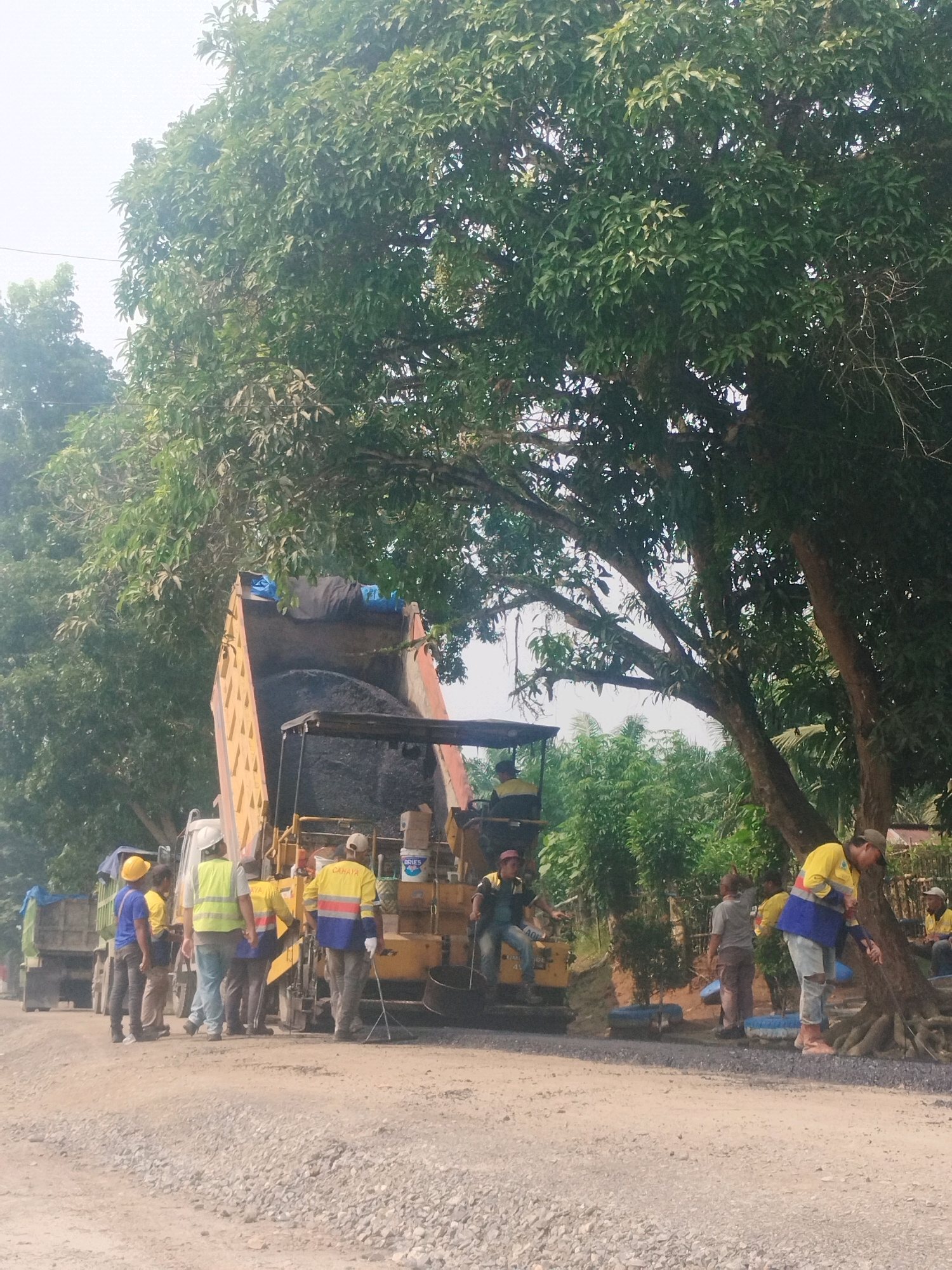 A group of construction workers are engaged in road paving in Pagarbosi, Indonesia. A dump truck is unloading asphalt into a paving machine, which is spreading the material on a gravel road. Several workers in high-visibility vests and hard hats are overseeing the operation, some are standing near the machinery, while others are observing the asphalt being laid. The scene is set outdoors, with large trees providing shade, and lush greenery in the background, indicating a tropical or subtropical environment. The sky is overcast.