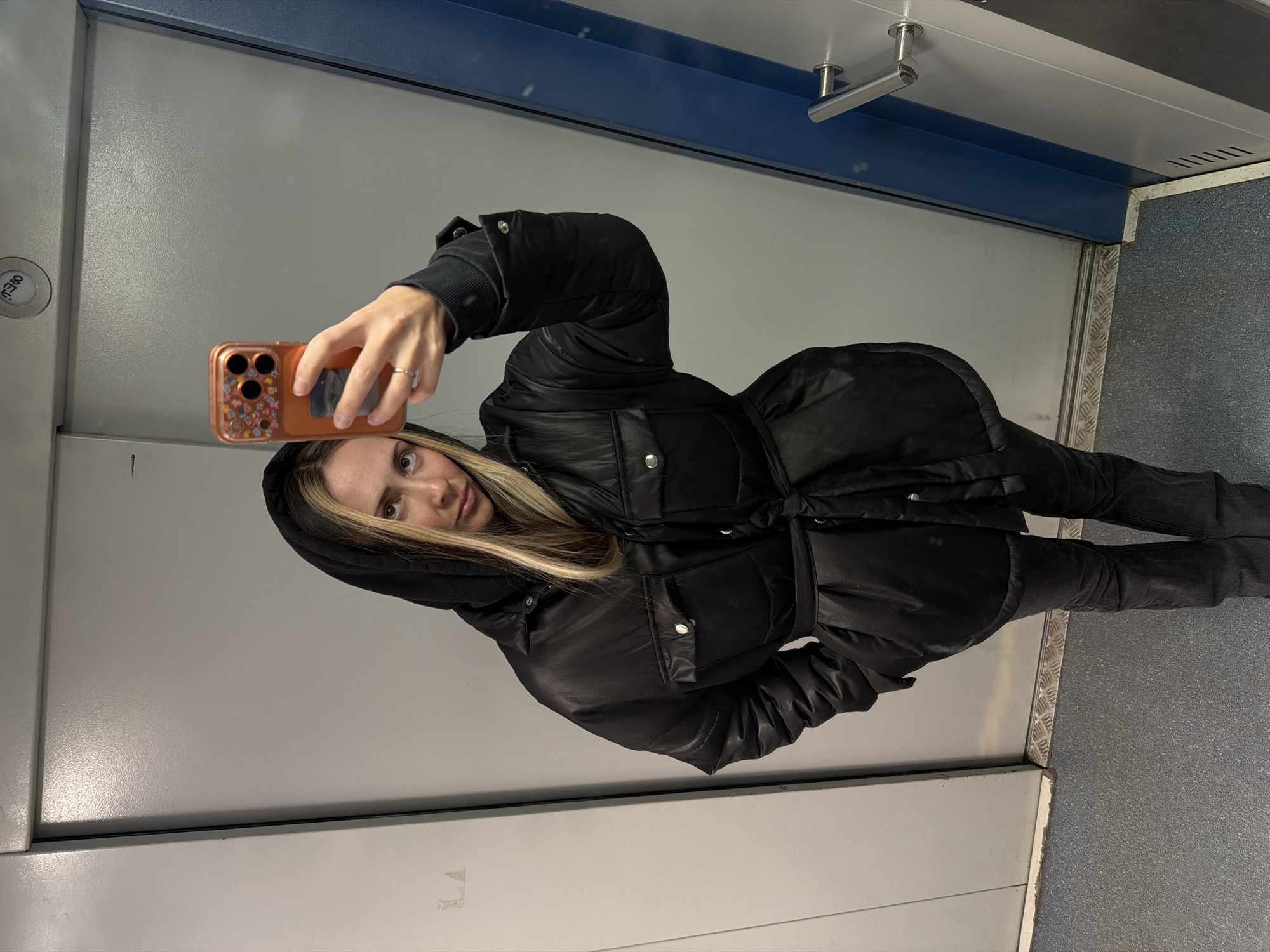 The image, rotated 90 degrees counter-clockwise, captures a woman taking a mirror selfie in an indoor setting. She looks directly at the camera with wide, light-colored eyes and a neutral expression. Her long, blonde hair, with darker roots, frames her face and is partly covered by a black hoodie.
The woman is wearing a black, belted puffer jacket with silver snap buttons on the pockets, along with dark trousers or jeans. Her right hand, adorned with a simple ring, holds an orange smartphone. The phone case features a floral pattern and a visible card slot on its back.
The background suggests a functional interior space, likely an elevator or a public corridor. The walls are light grey panels, featuring a distinct blue horizontal band along the upper section. A silver door handle is visible above this blue band on the right. The floor is covered in a textured grey material, with a silver diamond-plate trim running along the base of the wall. On the left wall, a round button displays partially obscured text that, when re-oriented, could read "OPEN" or "MENU". A small, dark ventilation grille is visible in the upper right corner. The lighting is uniform, characteristic of an interior environment. This scene is set in Khimki, Russia.