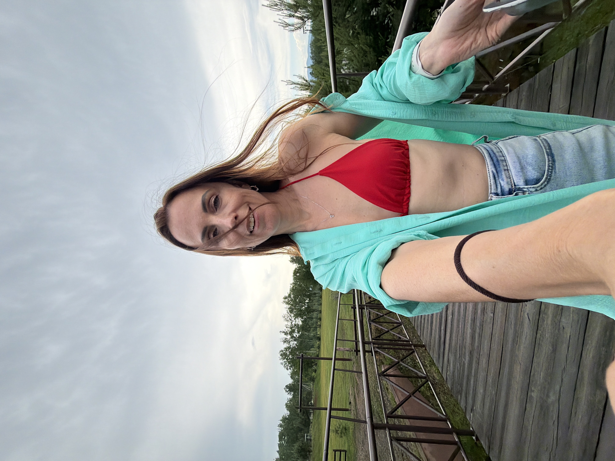A woman is pictured from the chest up, holding the camera as if taking a selfie. She is wearing a red bikini top and a turquoise open shirt, and light blue denim shorts. She has long brown hair that is blowing in the wind. Her left arm is extended towards the camera, and she is wearing a dark brown bracelet. The woman is standing on a wooden walkway that appears to be a bridge. Behind her, there is a grassy field with some wooden structures and trees. The sky is overcast with clouds. The location is identified as Birobidzhan, Russia.