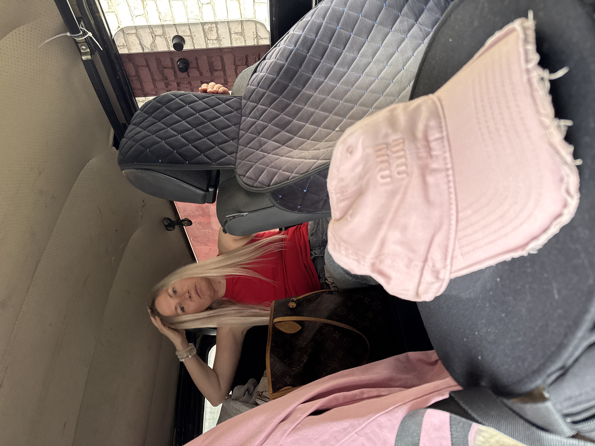 A blonde woman with long, wavy hair is lying on her side in the back of a vehicle. She is wearing a red tank top and blue jeans. Her right hand is resting on her head, and her left arm is extended out. She is looking towards the camera with a neutral expression.
In the background, there are several objects, including a quilted seat cover, a pink baseball cap with "MIU" embroidered on it, and a brown designer handbag. The interior of the vehicle appears to be well-maintained, with a light-colored headliner and dark-colored seats. The overall scene is casual and appears to be an indoor setting.
The image is shot from a slightly high angle, looking down into the back of the vehicle. The lighting is soft and even, suggesting it might be daytime. There are no specific clues about the weather or time of day. The location context provided is Karachayevsk, Russia.