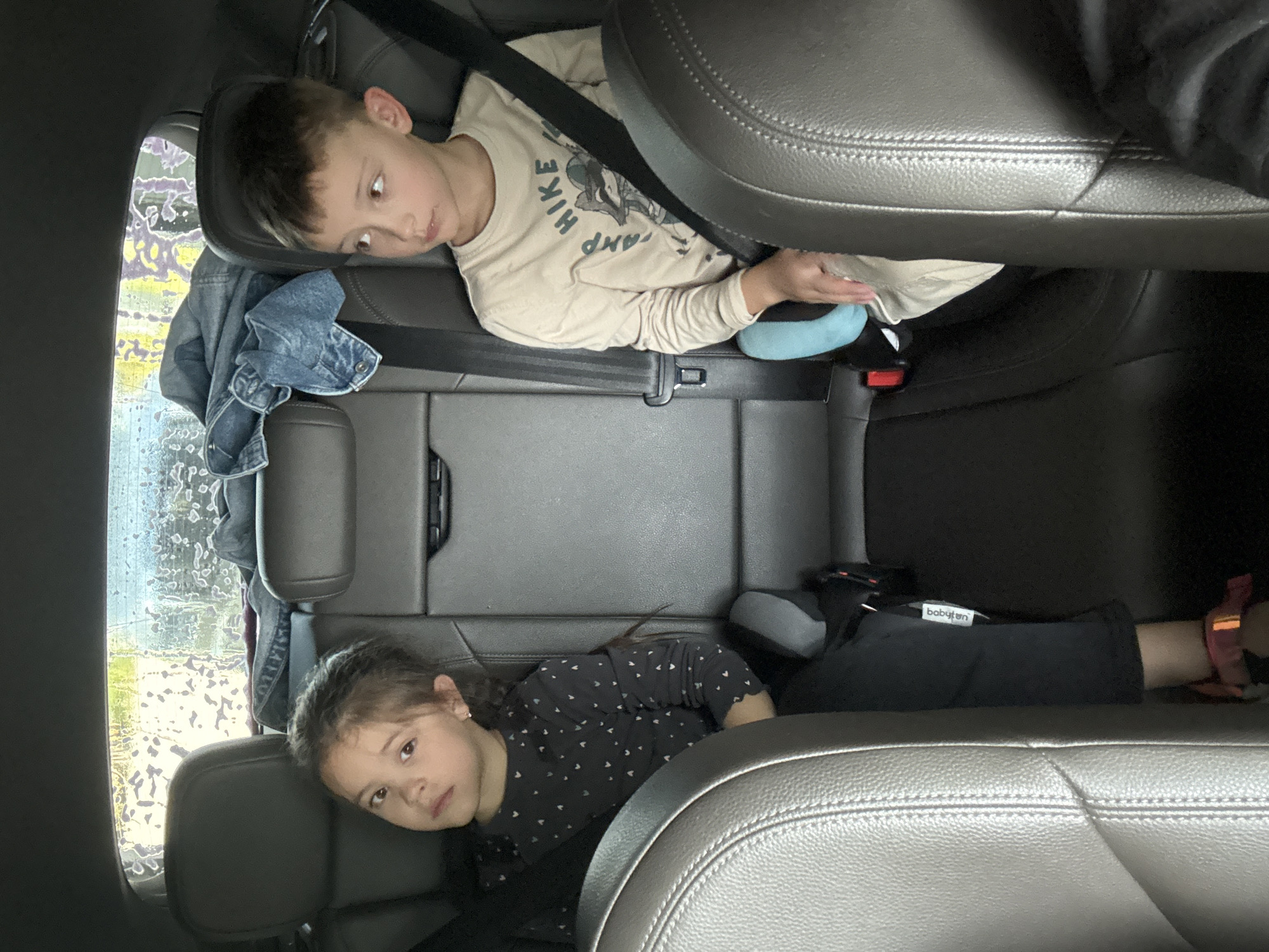 Two children are seated in the back of a vehicle. On the left, a young girl with dark hair pulled back sits facing forward, looking towards the camera. She is wearing a dark long-sleeved shirt with a pattern of small white hearts. She is secured by a seatbelt. Next to her on the right, a young boy with light brown hair sits facing forward, looking to his right. He is wearing a light-colored long-sleeved shirt with a graphic that appears to say "CAMP HIKE" and depicts a raccoon. He is also secured by a seatbelt. The interior of the vehicle appears to be leather. In the background, through the rear window, a blurry, abstract pattern of colors, possibly from a car wash or rain on the window, is visible. A folded denim jacket is also present on the rear seat behind the children. The image depicts a common scene of children traveling in a car. There is no information in the image to suggest a specific location within Votkinsk, Russia.