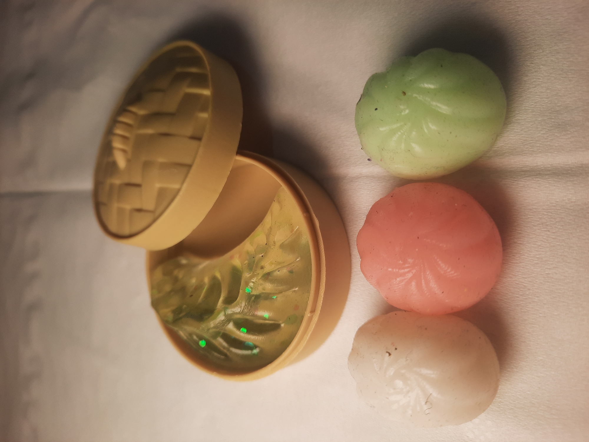 The image displays a close-up of three small, pastel-colored, swirled objects arranged in a row on a white, textured surface. From left to right, the objects are white, pink, and pale green. To the upper right of these objects, there is an open, light tan-colored container with a lid. The container has a woven pattern on the top of the lid. Inside the container is a translucent, pale green substance with small, green, sparkly flecks scattered throughout. One green fleck is prominently visible on the left side of the substance. The lighting in the image is soft, casting subtle shadows. The overall scene appears to be a still life composition of decorative items or toys. There is no discernible location in the city of North Jakarta, Indonesia, depicted in this image.