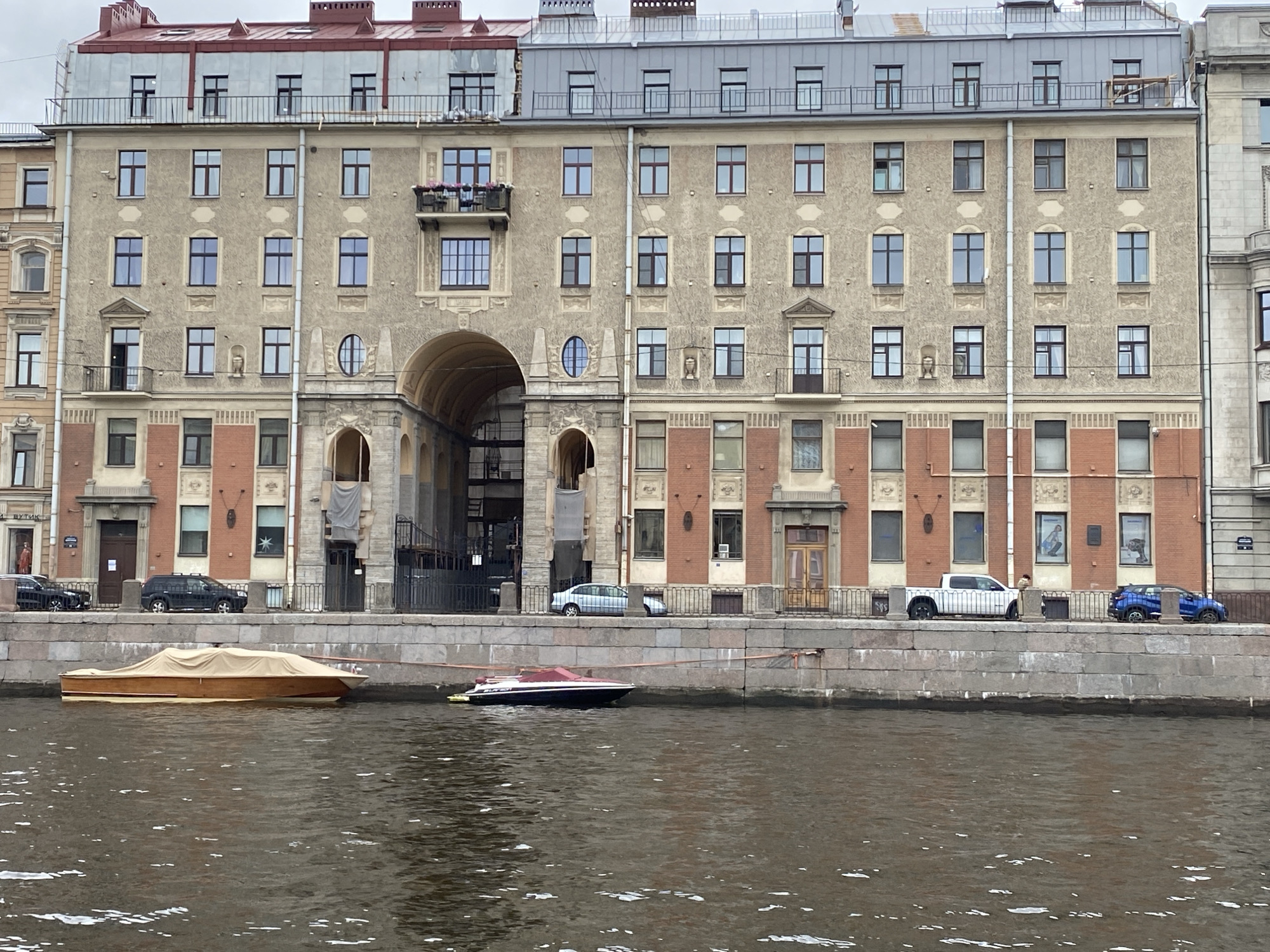 The image depicts a canal in Saint Petersburg, Russia, with a grand building lining its bank. The building exhibits a pale yellow facade, punctuated by numerous windows with dark frames. A prominent arched entryway suggests access to an inner courtyard or passage. The top of the building features a grey metal roof with visible structures and railings. Below the building, a stone embankment separates it from the canal. Two boats are visible on the water: a wooden boat covered with a tan tarp, and a sleek motorboat with a red cover. Several cars are parked along the embankment, including a black SUV, a silver sedan, a white pickup truck, and a blue hatchback. The sky is overcast, indicating a cloudy day.