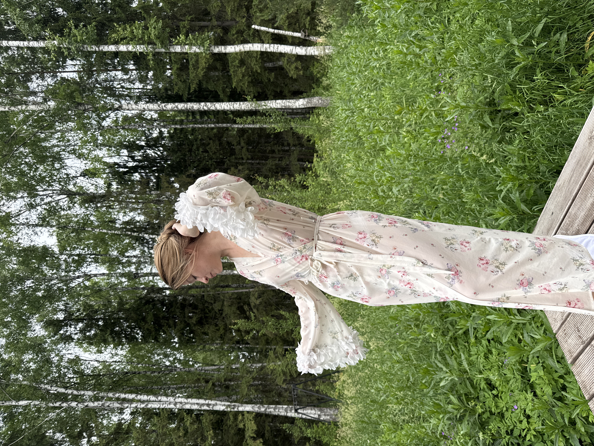 A woman stands on a wooden platform in a forest, wearing a sheer, long, floral-patterned robe with ruffled sleeves. The robe is light-colored with pink and blue flowers. Her hair is pulled back in a braid. She is looking down and to her left, with her right hand touching her head. The background is a dense forest with tall birch and evergreen trees, and a thick undergrowth of green plants and some small purple flowers. The sky is overcast. The image appears to be taken outdoors, in a natural setting likely outside the city of Bakhtiyary, Russia.