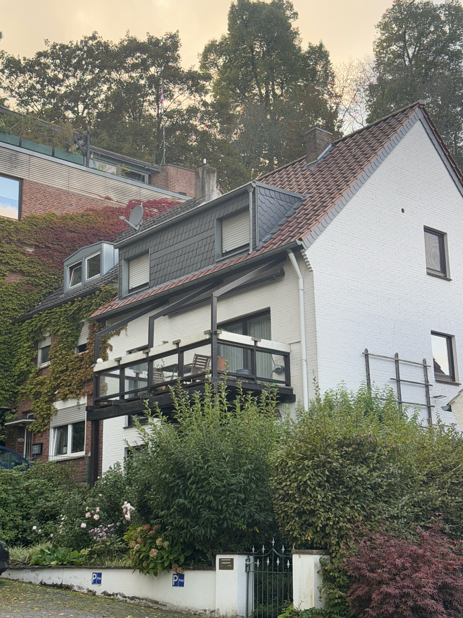 The image captures a multi-story residential building with a white brick facade on the right and a red brick facade covered in ivy on the left, situated in Cleves, Germany. The sky is overcast with a yellowish hue. The white building has a red tile roof and a black wooden balcony with glass railings, furnished with chairs. Several windows are visible on both buildings, some with shutters. A chimney is present on the white building's roof. Lush green foliage and bushes surround the base of the buildings, with a patch of pink and white flowers near a low white wall. A dark green metal fence with decorative elements is partially visible in the foreground, bordering a paved area. Two blue and white parking signs with the letter 'P' are mounted on the white wall. Trees with dense foliage are situated behind the buildings and on the slope above the red brick structure.