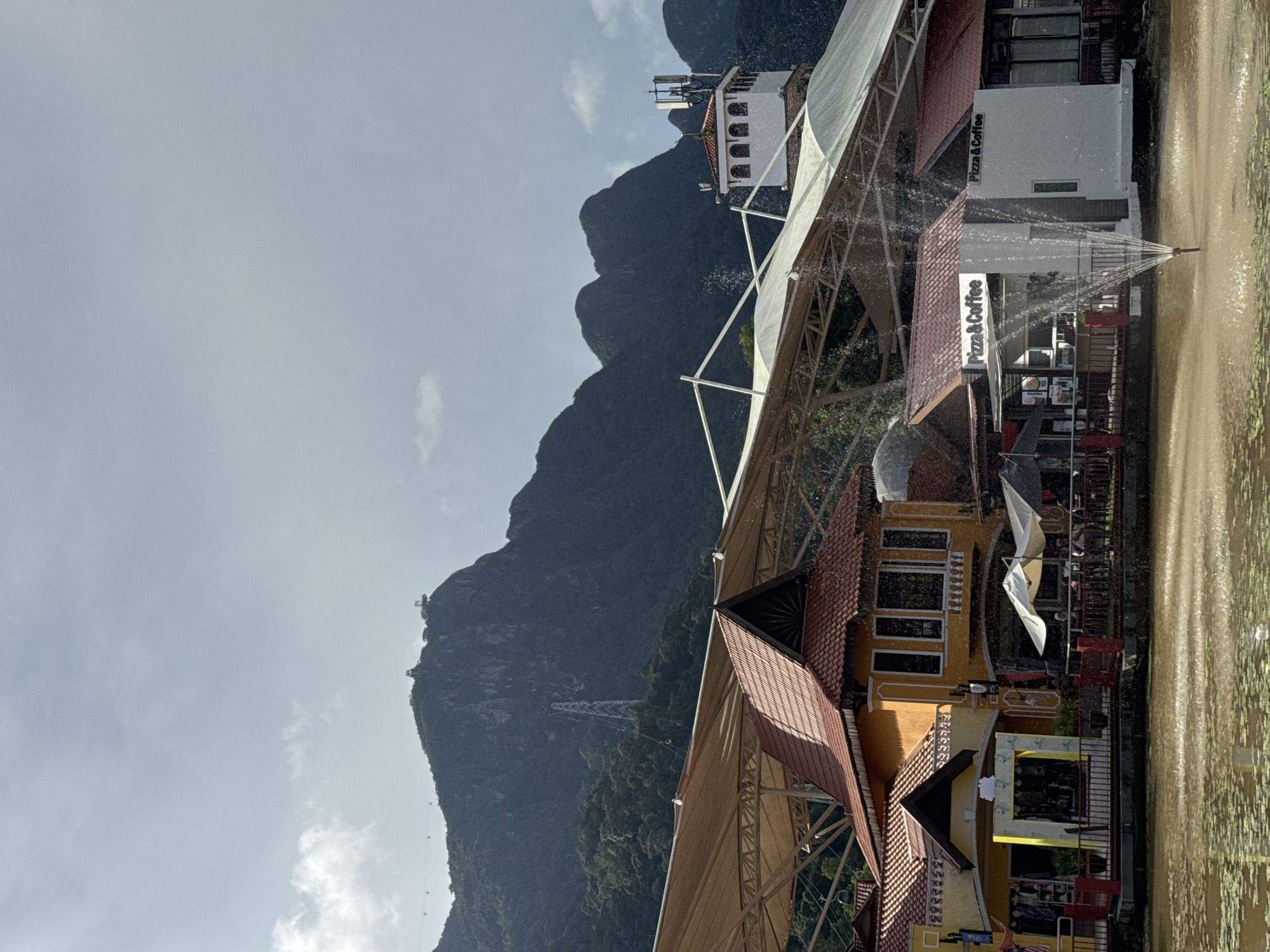 The image depicts a scenic view of the Oriental Village in Langkawi, Malaysia, a popular tourist attraction. In the foreground, a body of water with a fountain is visible, reflecting the overcast sky and surrounding structures. Several buildings with red-tiled roofs and Spanish-inspired architecture line the waterfront, some housing shops and cafes, with signage indicating "Pizza & Coffee." A large, beige canopy covers a section of the village, supported by a metal frame. In the background, a lush green mountain dominates the skyline, with a cable car system ascending its peak. A tall transmission tower is also visible on the mountainside. The overall scene is one of a tranquil, picturesque village nestled against a majestic natural backdrop.