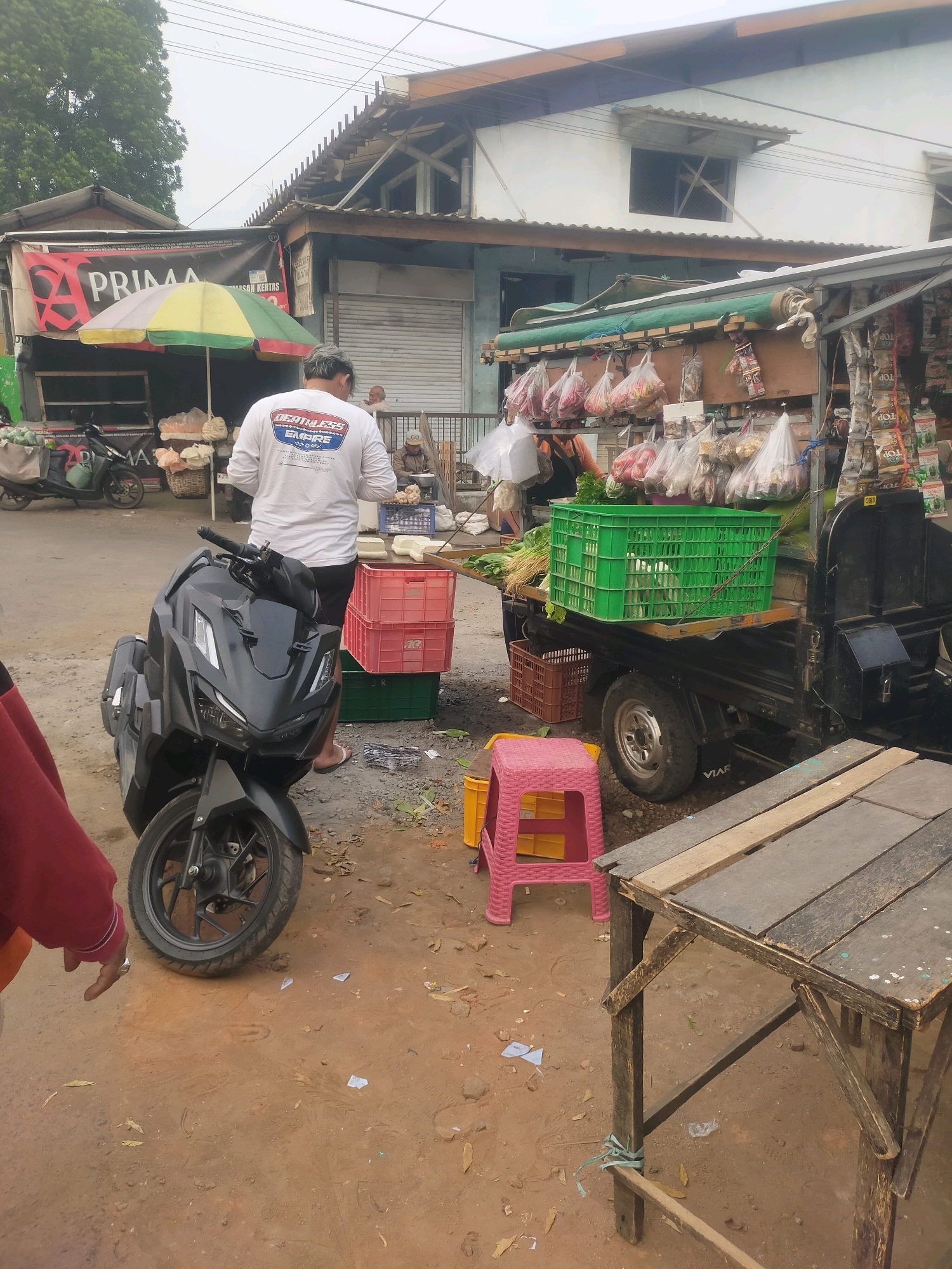 A street market scene unfolds in Bululawang, Indonesia, where a person stands near a black motorcycle. In the background, a vendor's cart is laden with various produce in plastic bags and green crates, alongside packaged goods. A banner reads "PRIMA" and an umbrella provides shade. Other individuals are visible in the distance, interacting with the market. The ground is dirt, with scattered leaves and debris. The environment appears to be an outdoor, open-air market.