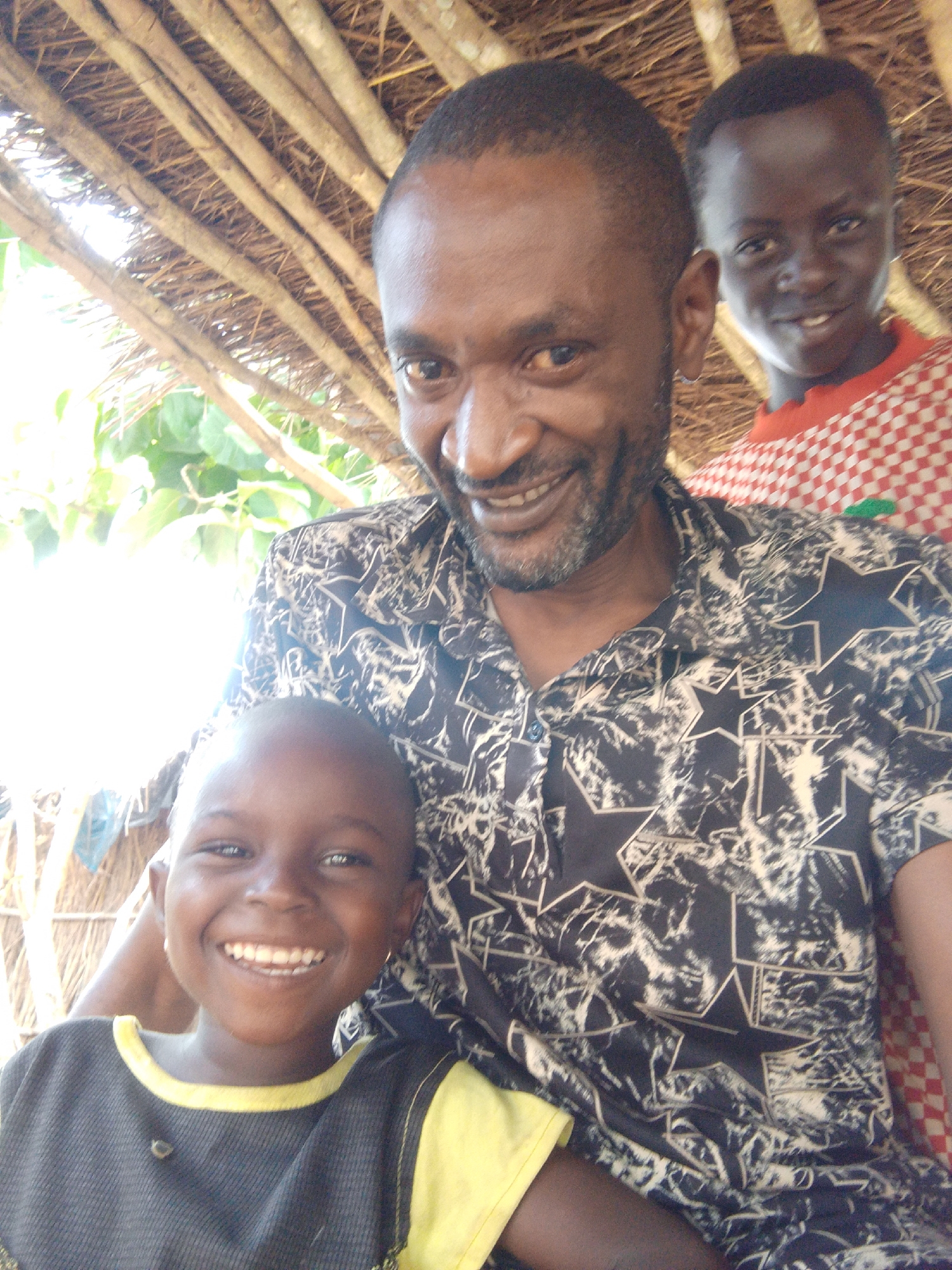 In Otulu, Nigeria, three individuals are captured in a close-up photograph. The foreground features a young child, their face beaming with a wide smile, wearing a dark t-shirt with a bright yellow collar. Behind them, an adult man, with a beard and a patterned shirt, smiles broadly, his eyes visible. Another young person is visible in the background, also smiling. The backdrop suggests a rustic setting, with a thatched roof structure overhead and foliage visible. The lighting is bright, with some areas appearing overexposed.
