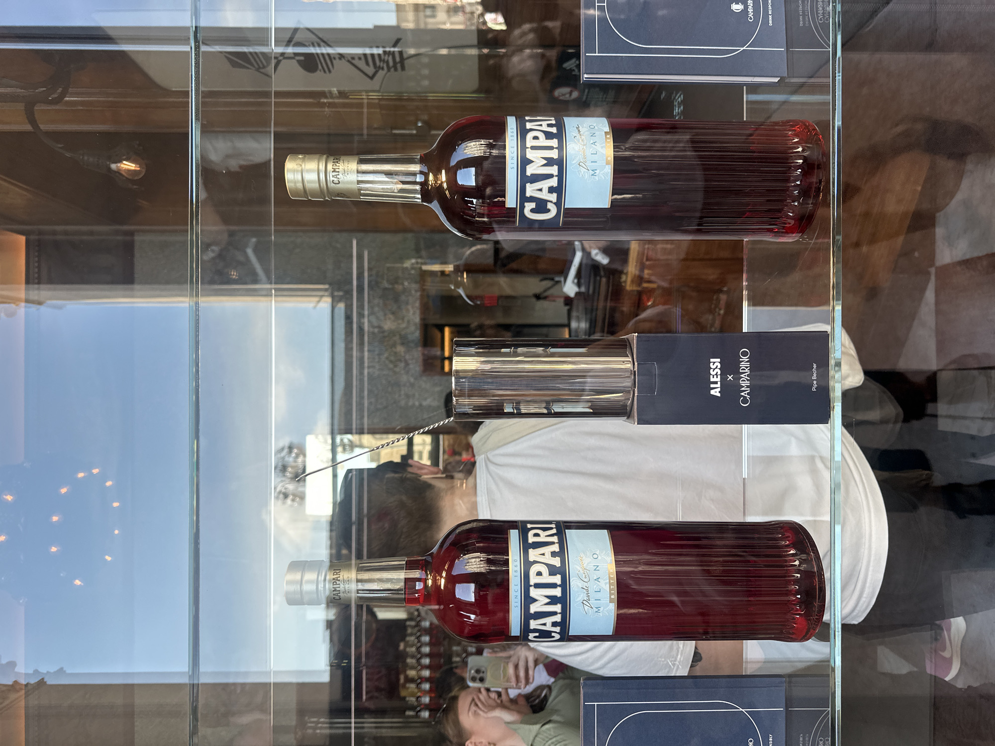 The image depicts a storefront display at Camparino in Galleria, Milan, Italy, viewed from the exterior through a large glass window. Two prominent, tall bottles of Campari Bitter are centrally positioned on a clear glass shelf. Each bottle contains a dark red liquid, features a gold-colored cap, and bears a label displaying "CAMPARI," "SINCE 1860," "Davide Campari," and "MILANO BITTER." The lower section of both bottles is vertically fluted.
Between the two Campari bottles, a cylindrical silver object, identified as an "ALESSI x CAMPARINO Pipa Becher," is displayed within its black packaging box. Portions of dark blue boxes, consistent with product packaging, flank the display items on the left and right sides of the shelf.
The clear glass window produces significant reflections of both the exterior environment and the interior of the establishment. A bright blue sky is prominently reflected above. Reflected against the sky is a distinct circular arrangement of approximately ten warm-toned, glowing light bulbs, characteristic of the interior ceiling lighting of the Galleria.
Multiple individuals are visible in the reflections. On the left, a woman with light brown hair, wearing a green top, is reflected holding a light-colored smartphone towards the display, appearing to photograph or video the items. In the center, the back of a person wearing a white t-shirt and dark trousers is reflected; a purple sneaker is visible on their left foot. Fainter reflections of additional individuals are discernible further in the background.
Behind the reflections of the foreground subjects, the dimly lit interior of the establishment is visible through the glass, featuring dark wooden paneling and shelves stocked with numerous bottles, consistent with a bar or retail liquor store. Several individual light bulbs suspended on black cords are also reflected within this interior view. The overall scene is illuminated by ambient daylight.