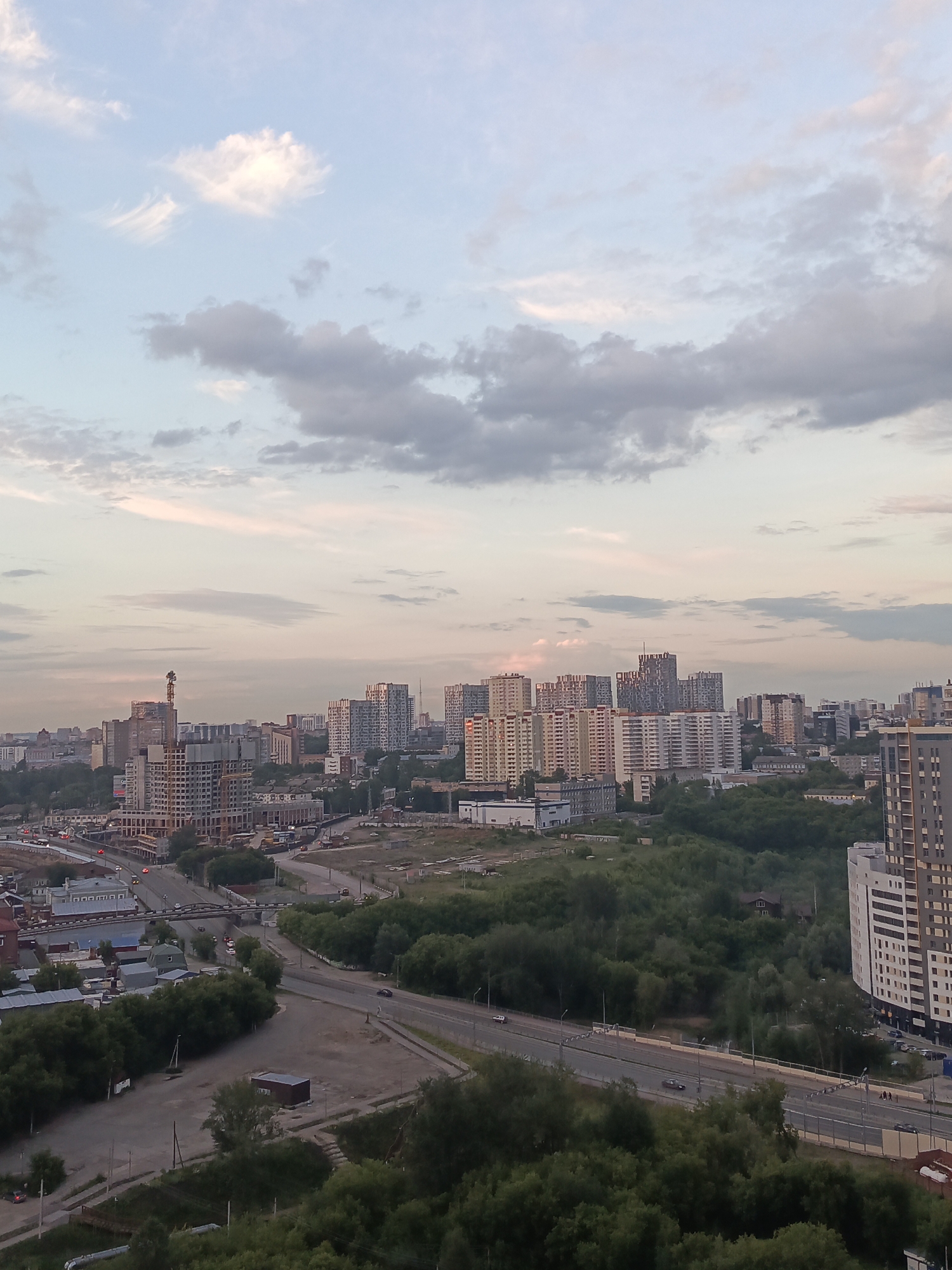 This is an aerial, long-shot photograph of a sprawling urban landscape in Perm, Russia, likely taken during dusk. The sky is a pale, soft blue with wispy clouds tinged with pink and orange, suggesting the sun is either rising or setting. The city below is a mix of modern high-rise apartment buildings, some under construction, and older, lower-rise structures. A prominent construction crane is visible on one of the unfinished buildings in the mid-ground.
Green trees and foliage are interspersed throughout the cityscape, creating a contrast between nature and the built environment. A multi-lane road, possibly a highway, runs through the scene, with a few cars visible on it. A bridge crosses over a road in the lower-left quadrant of the image. The overall impression is one of a developing city during a peaceful transition between day and night, with a serene and slightly muted color palette. There is no visible text in the image.