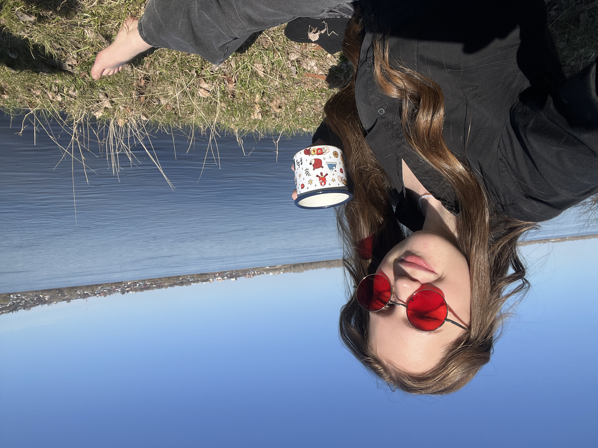 A young woman is reclined on grass near a body of water. She is wearing red sunglasses, a black collared shirt, and black pants. She is holding a white mug with cartoon illustrations. Her right foot is bare and rests on the grass. In the background is a body of water, a distant shoreline with buildings, and a blue sky. The location is Noviy, Russia.