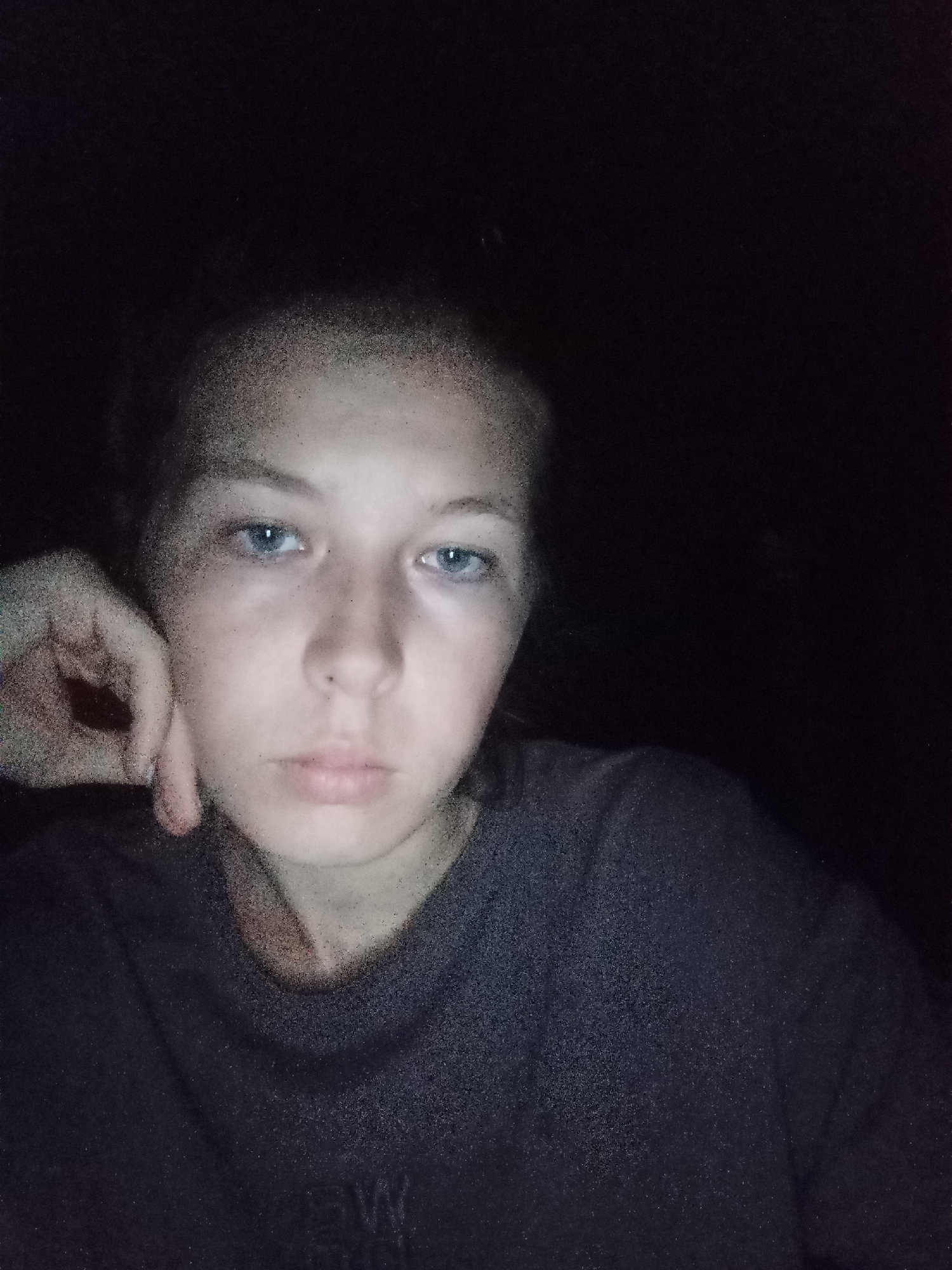 A selfie of a young, light-skinned person with blue eyes and short, dark hair, likely taken indoors at night given the dark background. They are resting their face on their hand and wearing a dark-colored t-shirt. The lower portion of the shirt has a very small, indistinct white mark. No indicators of location in Madaniyat (Ogonyok), Uzbekistan, are visible.