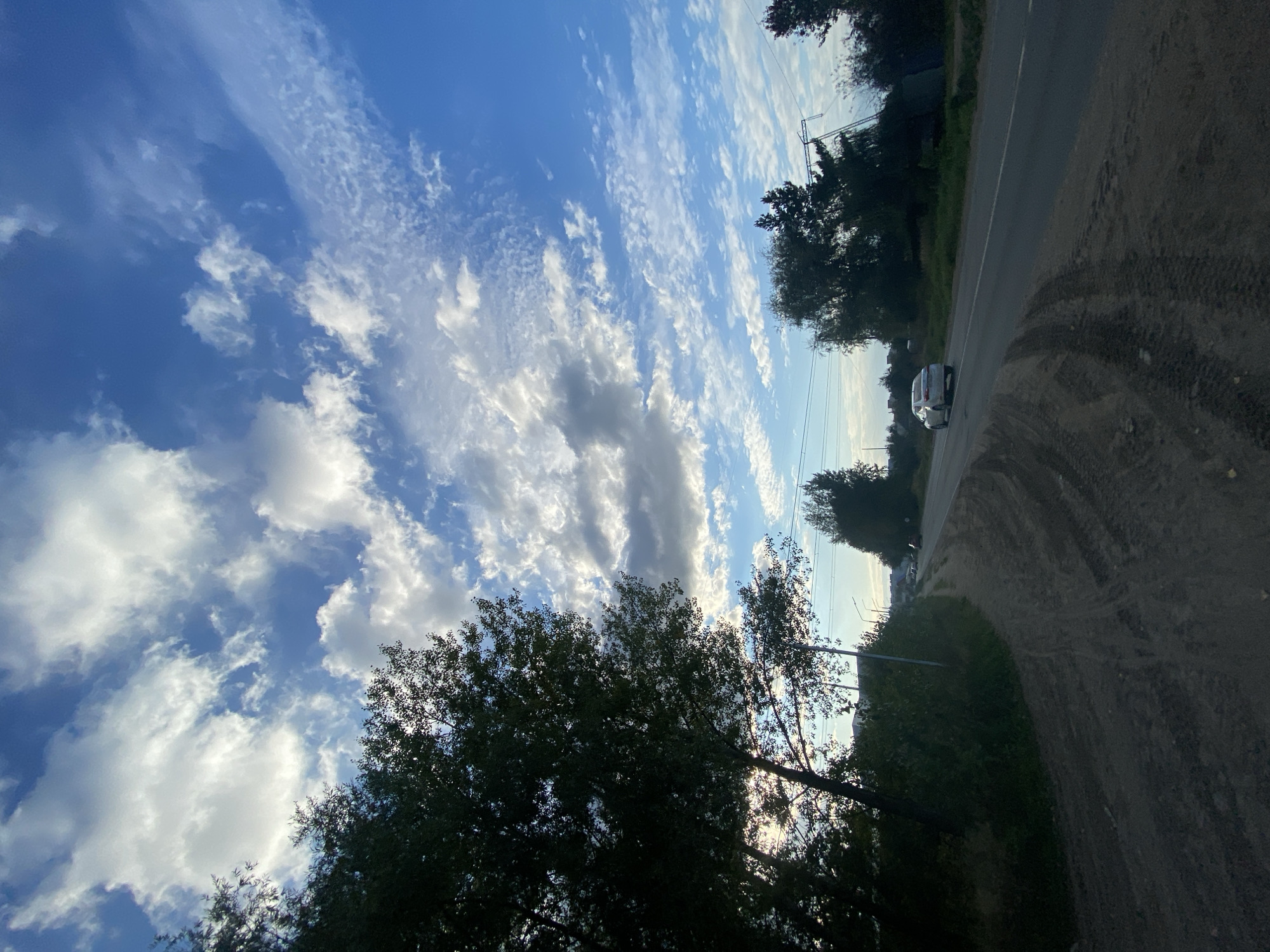 This is a vertical, outdoor, low-angle shot of a road in Chaikovsky, Russia. The sky is a vibrant blue with streaks of white and gray clouds, suggesting it's a partly cloudy day. Sunlight filters through the clouds and trees, creating a sense of depth and atmosphere, likely late afternoon. The road, a mix of asphalt and dirt, curves to the right, with tire tracks visible on the dirt shoulder. A white car is parked on the side of the road, and another car is visible further down. Lush green trees line the left side of the road, their branches reaching towards the sky. In the background, there are glimpses of what appear to be buildings and utility poles, suggesting a semi-urban or rural setting. The overall mood is peaceful and serene, with a hint of the outdoors and the open road. There is no visible text in the image.