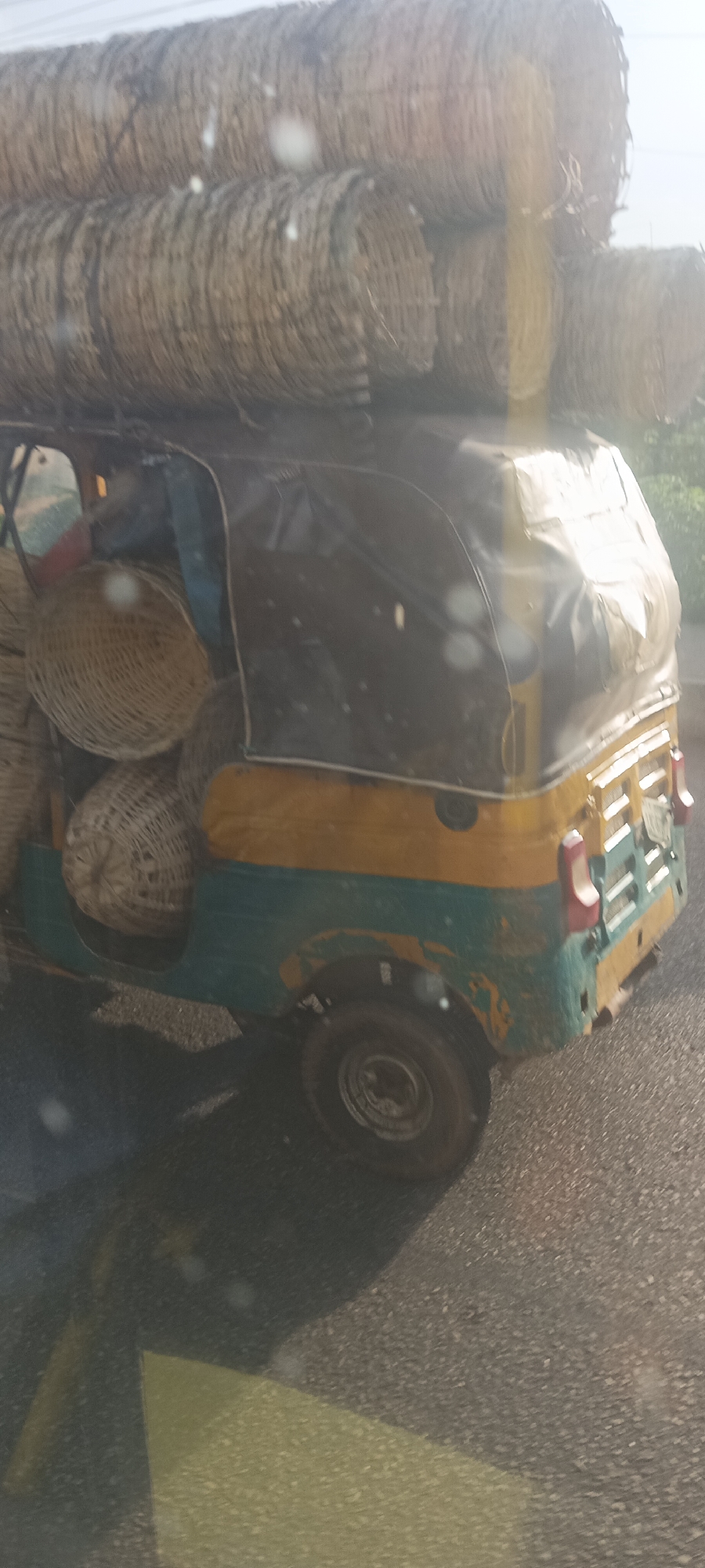 The image displays a heavily laden three-wheeled auto-rickshaw on a road, likely in Gura Top, Nigeria. The vehicle, painted in a distinctive combination of green and yellow/orange with a black fabric canopy, is filled and piled high with woven baskets.
The primary subjects are the auto-rickshaw and its cargo. On the roof, numerous cylindrical bundles of what appear to be tightly woven baskets or basket-like materials are stacked vertically and horizontally, creating a substantial load that extends well above the vehicle's height. Inside the open rear compartment, large, round, woven baskets are visible, packed snugly together.
The activity depicted is the transportation of goods, specifically these baskets, by the auto-rickshaw. The vehicle itself shows signs of wear, with some peeling paint on its body. The setting appears to be a typical road, possibly a rural or semi-urban route, with an asphalt or gravel surface visible beneath the vehicle.
Notable details include the sheer volume of the cargo, suggesting either a market delivery or a substantial collection/distribution task. The lighting is bright and somewhat hazy, indicating it is daytime, possibly late morning or afternoon, with lens flares suggesting the photo was taken through a window or windshield. No discernible text is visible on the vehicle or cargo. The general appearance of the auto-rickshaw and the type of cargo are consistent with local commerce and transportation in many parts of Africa.