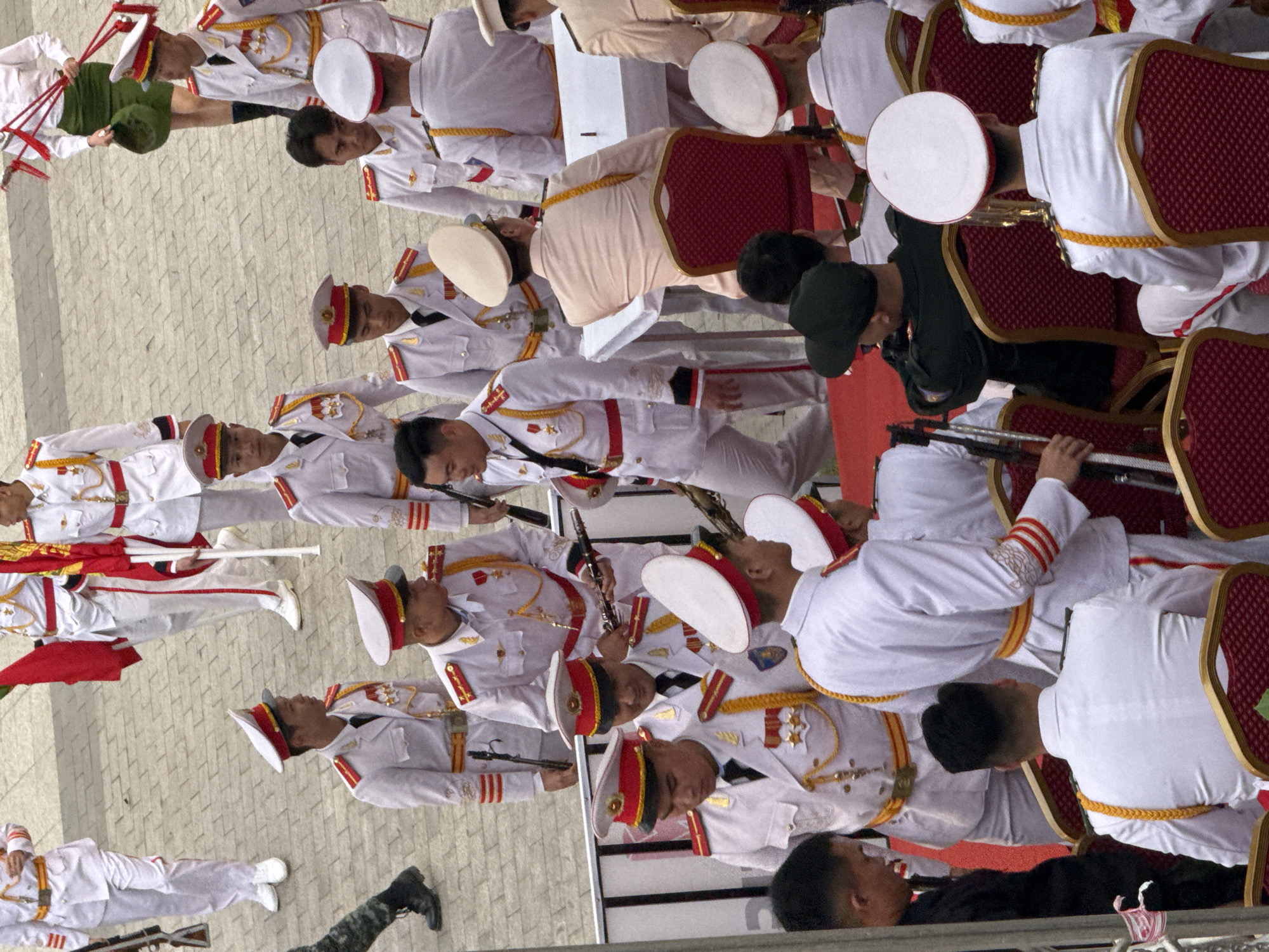 The image depicts a group of men in formal white military uniforms with red and gold accents, participating in an outdoor ceremony. The men are standing and some are seated on red chairs. They are wearing peaked caps and several are holding musical instruments, suggesting a band performance. The setting appears to be a paved plaza with a backdrop of buildings and flags, consistent with a public event. The question about the location being in Da Lat, Vietnam, cannot be confirmed from the visual information alone.