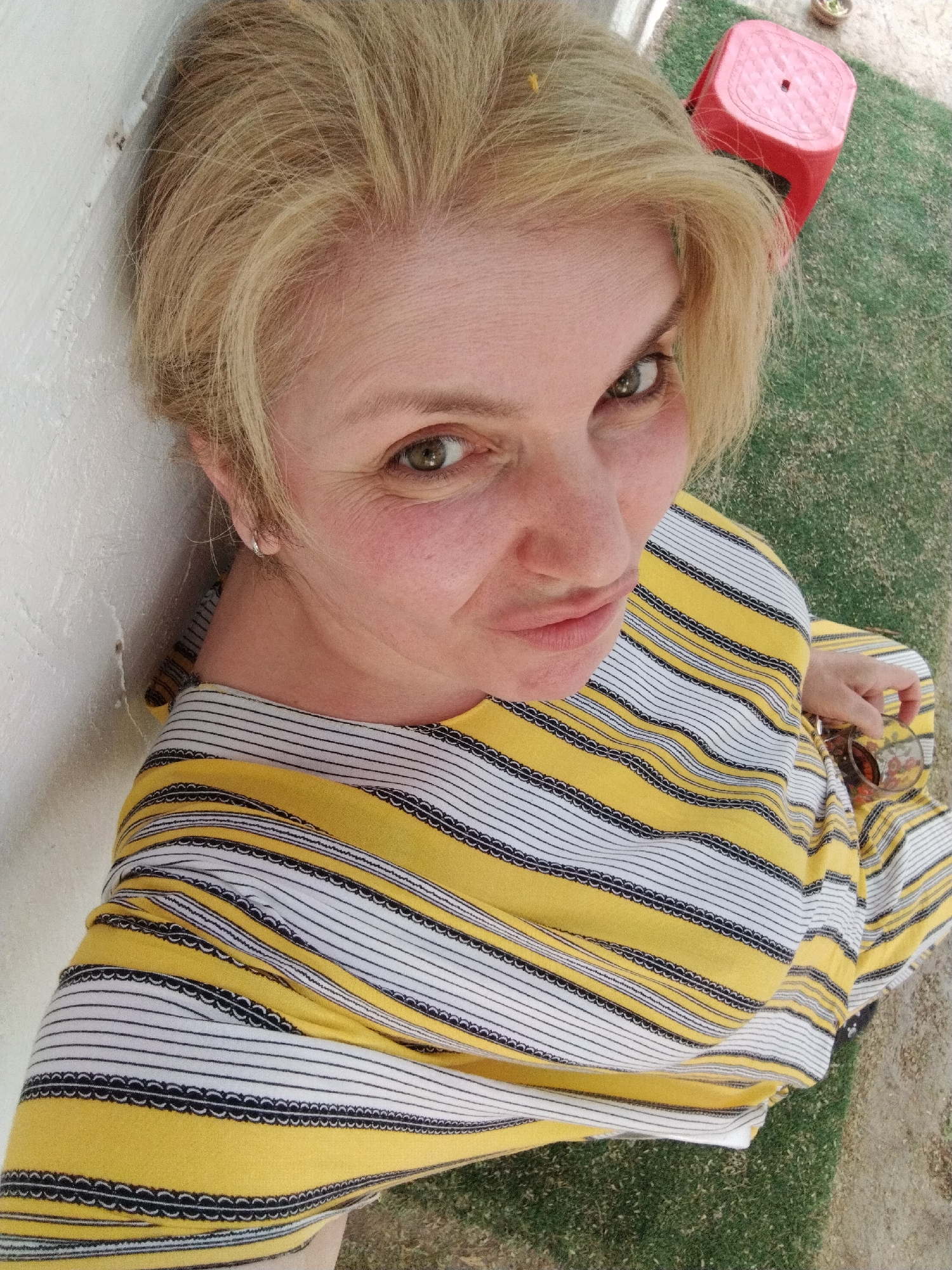 A blonde, light-skinned woman is pictured from a high-angle perspective against a white wall. She is wearing a yellow and white striped top with black detailing between the stripes. She is holding sunglasses in her right hand. A red plastic stool is visible in the upper right corner, resting on what appears to be artificial turf. The scene appears to be outdoors. There is no evidence in the image to suggest that the photo was taken in Al Mas'oudi, United Arab Emirates.