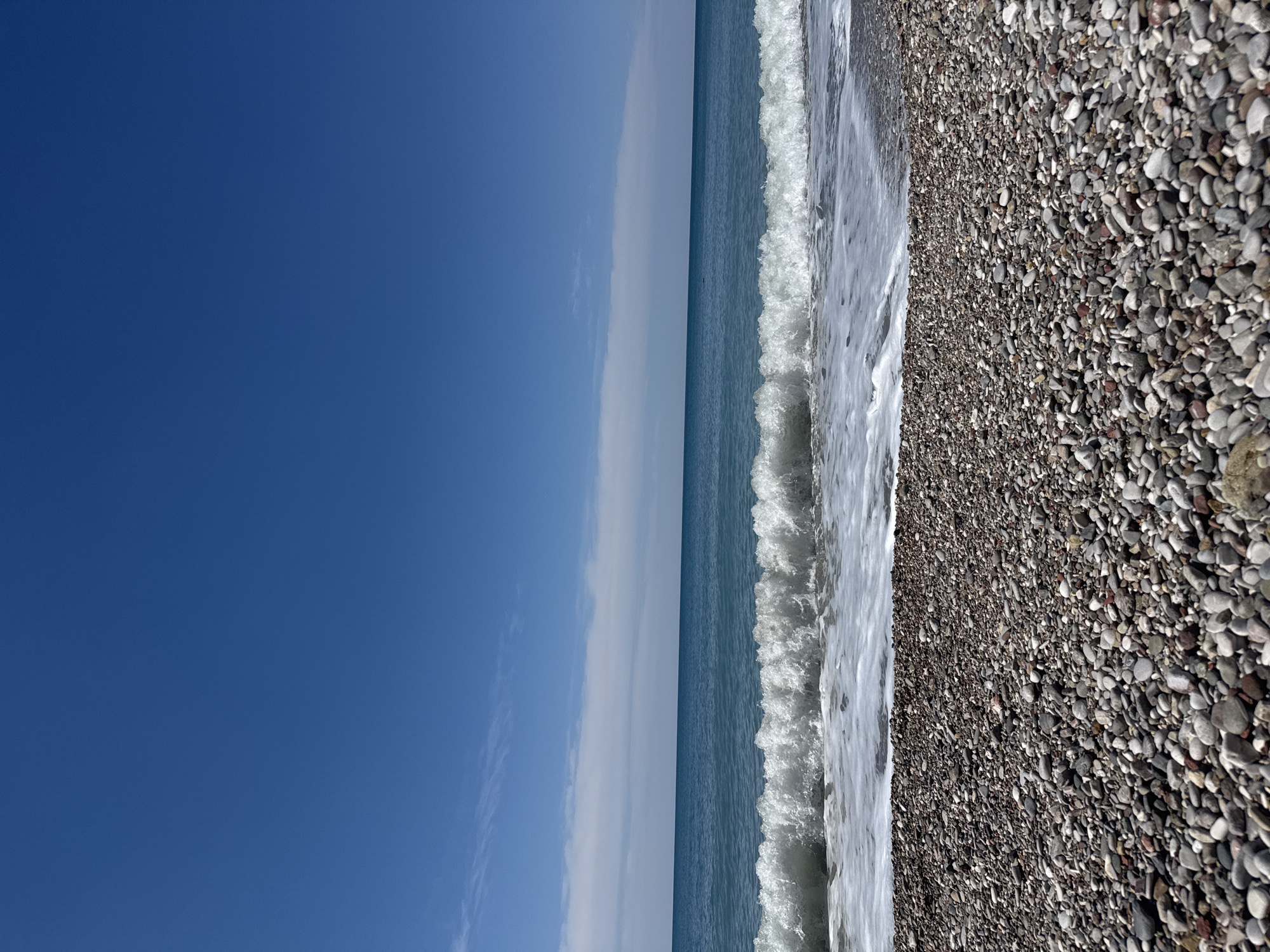 The image presents a seaside view, showing a beach with small stones along the coast. The water is blue, with waves reaching the shore. The sky is predominantly blue with some scattered clouds near the horizon. The image is a view of the coastline in Bar, Montenegro.