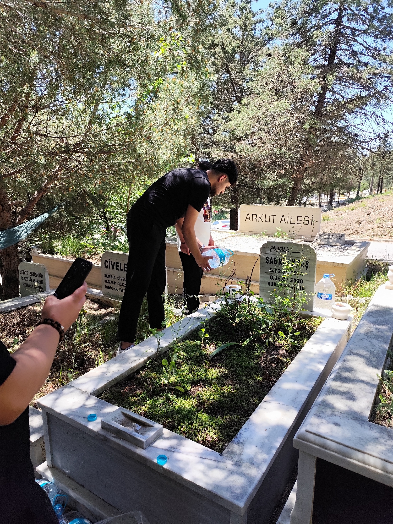 This image shows two people tending to a grave in a cemetery. One person, dressed in black, is pouring water from a large bottle onto the grave. Another person is standing next to them, partially visible. In the foreground, a person is holding a black smartphone, possibly taking a picture or video.
The grave is enclosed by a white marble structure. Various bottles of water and blue bottle caps are visible around the grave. The gravestone reads "Sabri Kayaş," with dates indicating a death in 2020. Another visible gravestone reads "Arkut Ailesi."
The scene is partially shaded by trees, suggesting a natural environment. Based on the other visible gravestones "Hediye Sayginsoy" and "Civele", this cemetery is located in Sarıyer, Turkey. The overall environment appears to be a sunny day.