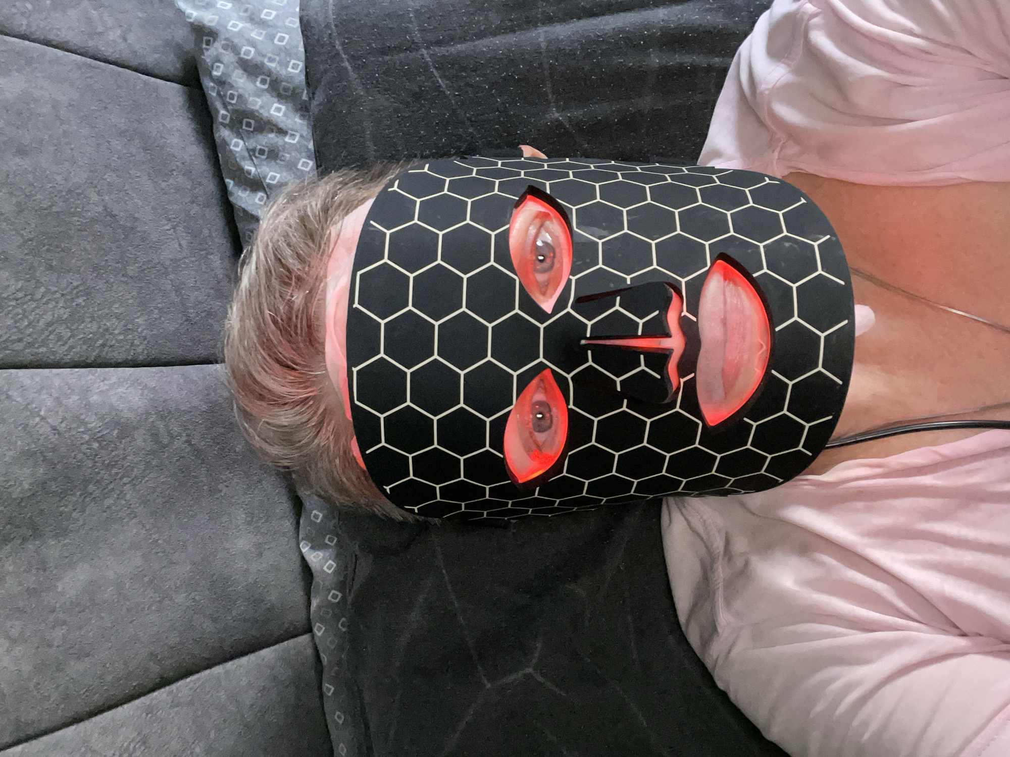 A person wearing a black LED face mask with a hexagonal pattern lies on a bed. The mask covers their entire face, with openings for the eyes, nose, and mouth, through which red light emanates. The person's hair, appearing to be a light brown or blonde, is visible around the edges of the mask. They are wearing a light pink shirt, and a thin silver chain is visible around their neck. The background consists of a grey upholstered headboard and pillows, with a dark gray blanket or sheet visible on the bed. The scene appears to be indoors, likely in a bedroom.