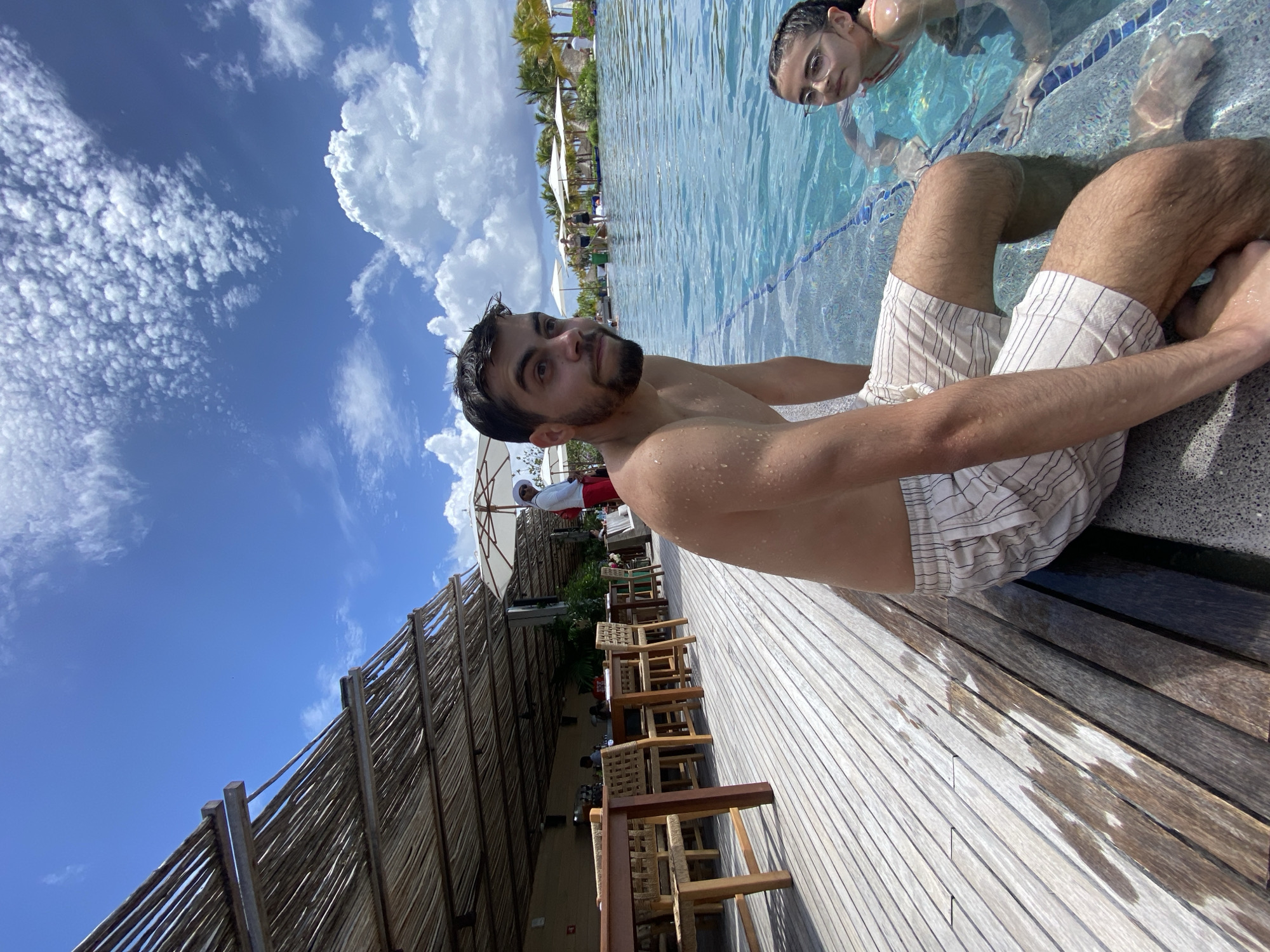 The image shows a man sitting on the edge of a pool in Marina Maroma, Mexico. He has dark hair and a beard and is wearing striped shorts. His torso is bare. Another person is in the pool behind him. The area beside the pool features a wooden deck with seating and a thatched roof structure. The sky is blue with white clouds.