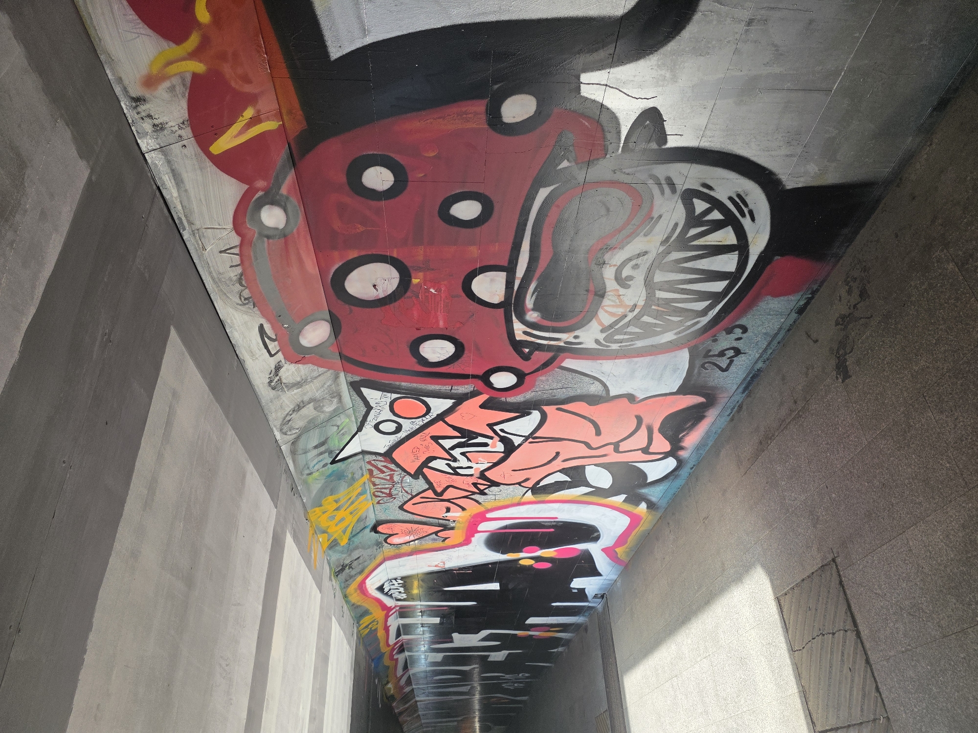 The image shows graffiti art on the underside of a concrete overpass or bridge. The art includes several cartoonish characters and abstract shapes in vivid colors, including red, black, white, yellow, pink, and blue. One prominent figure is a red shape with white circles. Another has a large, open mouth with sharp teeth. Other elements include stylized lettering and graphic designs. The structure is a gray concrete, and the perspective is looking upwards into the overpass, giving a sense of depth and dimension. Light is visible at the end of the tunnel. The city and location of the graffiti are not clear based on the information given in the image.