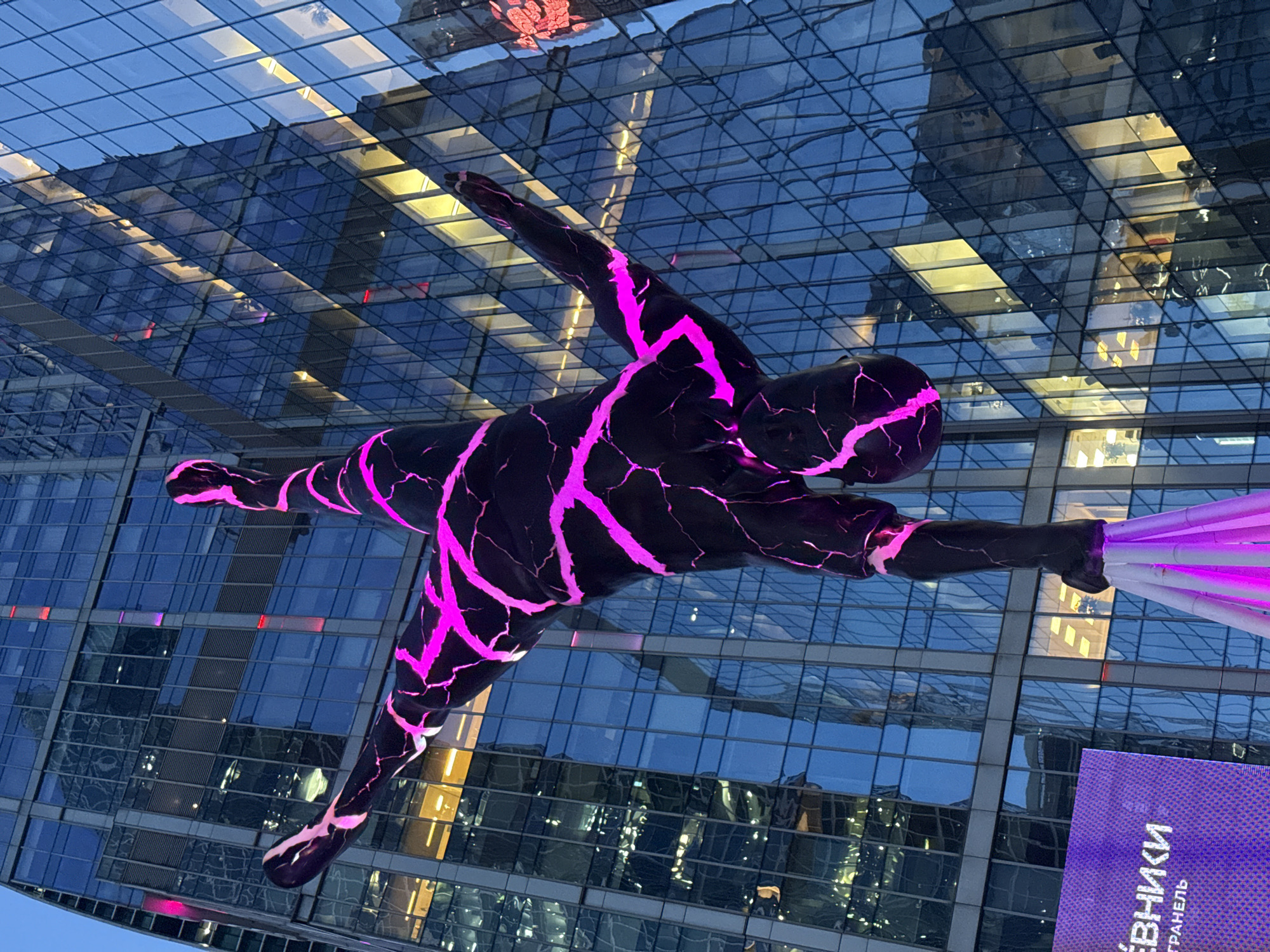 An outdoor, low-angle, full shot captures a colossal, black sculpture of a human figure, cracked with glowing pink lines, suspended upside down. The sculpture is positioned in front of a modern, glass-facade skyscraper in Moscow, Russia. The building's windows reflect the ambient light, and some windows show the glow of interior lighting, suggesting it is evening. In the lower left of the frame, a purple LED billboard displays Cyrillic text, indicating its likely location within a public or commercial space. The sky above is dark, hinting at nighttime or twilight. The sculpture's pose is dynamic, with limbs extended, appearing to be mid-movement. The overall scene is illuminated by the sculpture's internal lighting and the building's lights, creating a dramatic and somewhat surreal atmosphere.