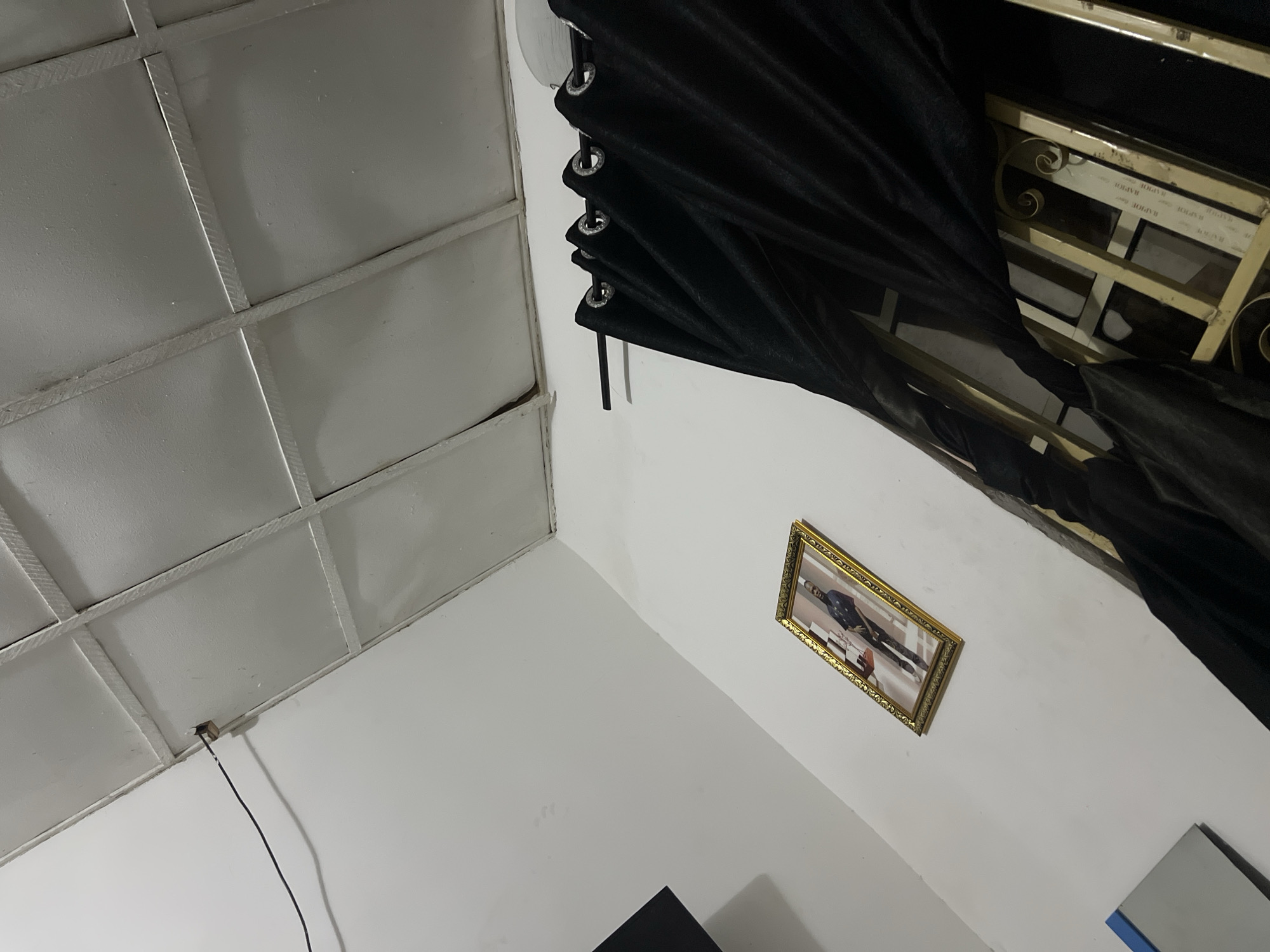 This is a close-up, eye-level, interior shot of a room with white walls and a white, grid-patterned ceiling. A black cord is visible running from a small wooden anchor on the ceiling towards the left side of the frame. Below the ceiling, on the left wall, a framed portrait of a man in a blue uniform is hanging. To the right of the portrait, a dark black curtain with metal grommets hangs from a dark rod, partially obscuring a window with a gold-colored ornate grill. The overall impression is one of a simple, domestic interior space. This image was captured in the city of Howayi, Nigeria.