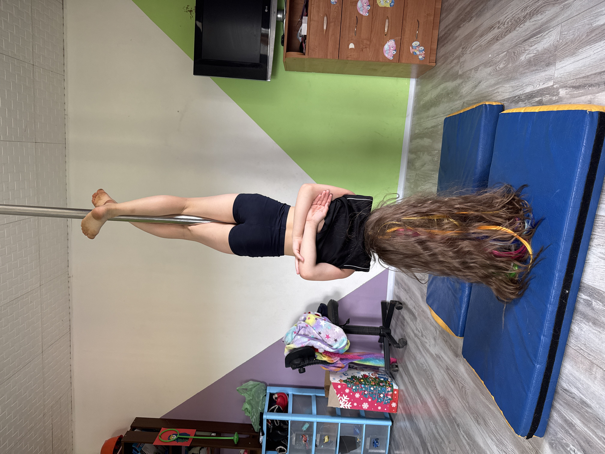 A young girl with long, multi-colored hair is upside down, suspended horizontally on a silver pole. She is wearing black shorts and a black t-shirt. Her arms are behind her back, hands clasped. Her legs are straight, with her feet pointed. The scene appears to be indoors, likely a dance or gymnastics studio, given the presence of blue crash mats on the floor. The walls are painted in distinct sections of white, light green, and light purple, creating a geometric pattern. To the left, a white tiled wall is visible. In the background, a wooden cabinet with decorative knobs and a television screen mounted on the wall are present. A rolling office chair with a pile of colorful blankets and clothes on it is positioned near the girl. Further to the right, more crash mats are visible, and the floor has a wood-like laminate finish. A blue storage unit with various items in its compartments is also in the background. There is no indication of the time of day or weather. No visible text is present in the image.