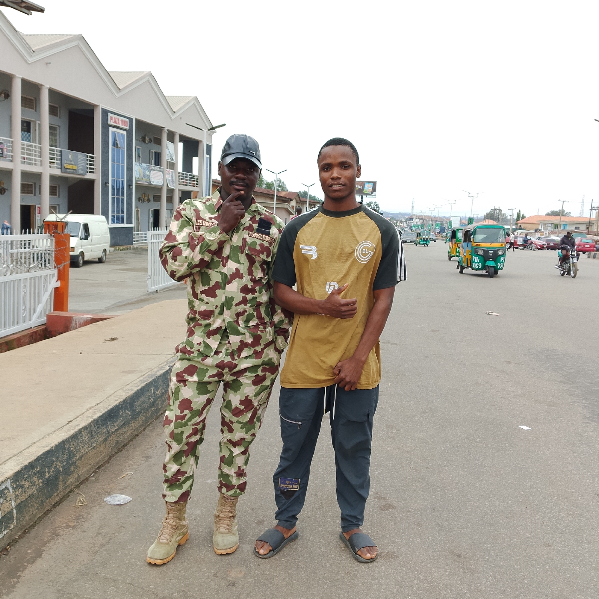 Two young men are standing on a paved road in what appears to be a town or city. The man on the left is wearing a camouflage military uniform with a cap. He has his right hand raised to his chin in a thoughtful pose. The man on the right is wearing a tan t-shirt with dark sleeves and dark pants. He is looking towards the camera with a slight smile.
In the background, there are buildings, including one with a sign that reads "PLAZA 1080". There are also several vehicles on the road, including a white van, a few motorized rickshaws (also known as 'keke napep' or 'marwa'), cars, and a motorcycle. The sky is overcast, suggesting it might be a cloudy day or early morning/late afternoon. The overall scene depicts a casual moment on a street, possibly in Nigeria, given the text on the military uniform "NIGERIAN ARMY".