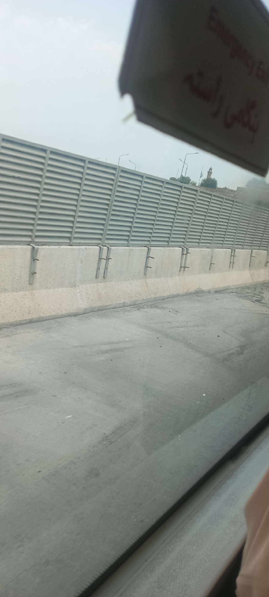 The image captures a view from a vehicle, looking towards a grey, slatted sound barrier wall constructed along a road. The wall is supported by concrete pillars. In the upper right corner of the image, a dark sign with English and Urdu text is partially visible. The English text reads "Emergency Exit." The Urdu text, written in a script with a reddish hue, appears to be "نگاہی راستہ" which translates to "Gazing Path" or "Viewing Path." In the background, a minaret of a mosque is visible above the sound barrier, suggesting an urban or semi-urban environment. The road in the foreground is grey and appears somewhat worn. The sky is overcast and hazy. There are no people or vehicles clearly depicted in the scene. The overall impression is of a functional infrastructure element within a Pakistani city.