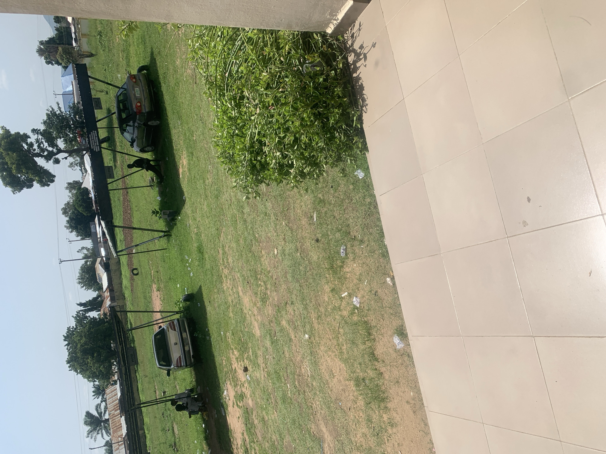 The image depicts an outdoor scene, likely in Jalingo, Nigeria, with a focus on two parked cars under a covered shelter. In the foreground, a tiled walkway with a beige, square tile pattern is visible. To the right, a dense, green bush is growing. The middle ground shows a grassy area with scattered debris, including plastic bottles and wrappers.
One car, a silver sedan, is parked under the shelter on the left, with two individuals sitting nearby. Another car, a dark colored sedan, is parked under the same shelter further to the right, with one person sitting beside it. The shelter appears to be constructed with metal poles and a dark, corrugated roof. Behind the shelter, there are various buildings with corrugated metal roofs, some appearing to be stalls or small structures. Palm trees and other vegetation are visible in the background, along with utility poles and overhead wires, suggesting an urban or semi-urban environment. The sky is clear and blue, indicating a sunny day.