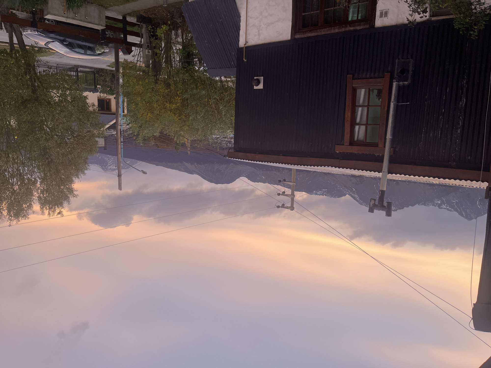 The image presents a landscape in Ushuaia, Argentina. Snow-capped mountains form a backdrop under a sky transitioning from sunset hues to a purple dusk. In the foreground is the corner of a dark, corrugated building with a window. Utility poles and power lines traverse the sky. To the left, hints of urban life appear, including trees, a residential street with a car, and adjacent buildings. The scene captures a blend of natural and urban elements typical of Ushuaia's setting.