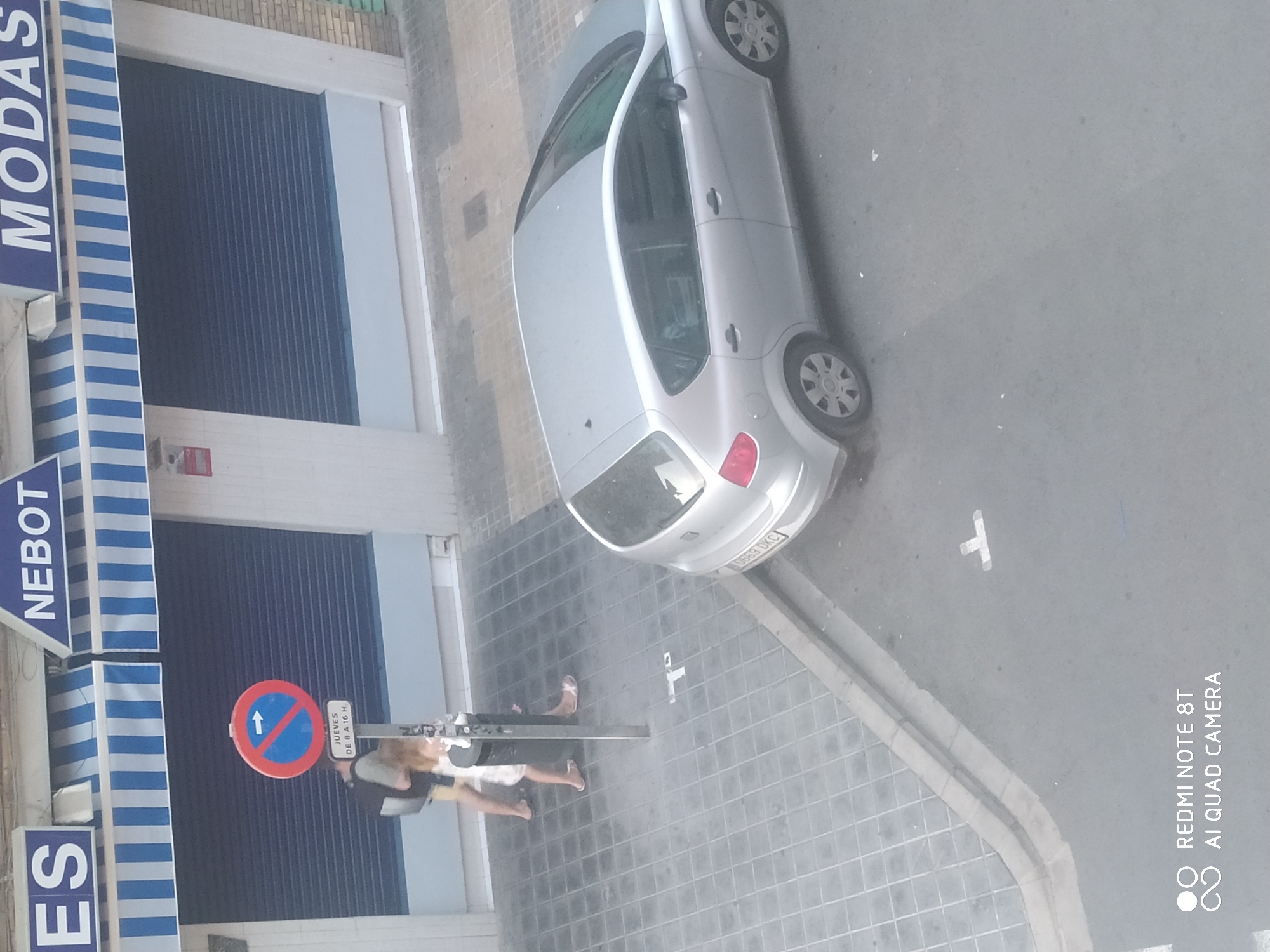 This is an eye-level, slightly angled, medium shot of a street scene. In the background, a building with blue and white striped awnings has "ES," "NEBOT," and "MODAS" written in large blue letters across its facade. The building has closed, dark blue metal shutters. To the left, a "no parking" sign with a white arrow pointing right is mounted on a pole. Below it, a white sign reads "JUEVES DE 8 A 16 H."
In the middle ground, a silver hatchback car is parked on the street. Two individuals are walking on the sidewalk. One, wearing a dark t-shirt and shorts, walks away from the camera with a backpack. The other, wearing a white dress, walks in the same direction, slightly ahead of the first person. Both are wearing sandals.
The foreground shows a paved sidewalk and a portion of the street. The sidewalk is made of square tiles. A curb separates the sidewalk from the asphalt street. White markings are visible on the street, one of which appears to be a 'T' shape. The bottom left corner of the image shows a watermark with "REDMI NOTE 8T" and "AI QUAD CAMERA." The overall lighting suggests it is daytime.